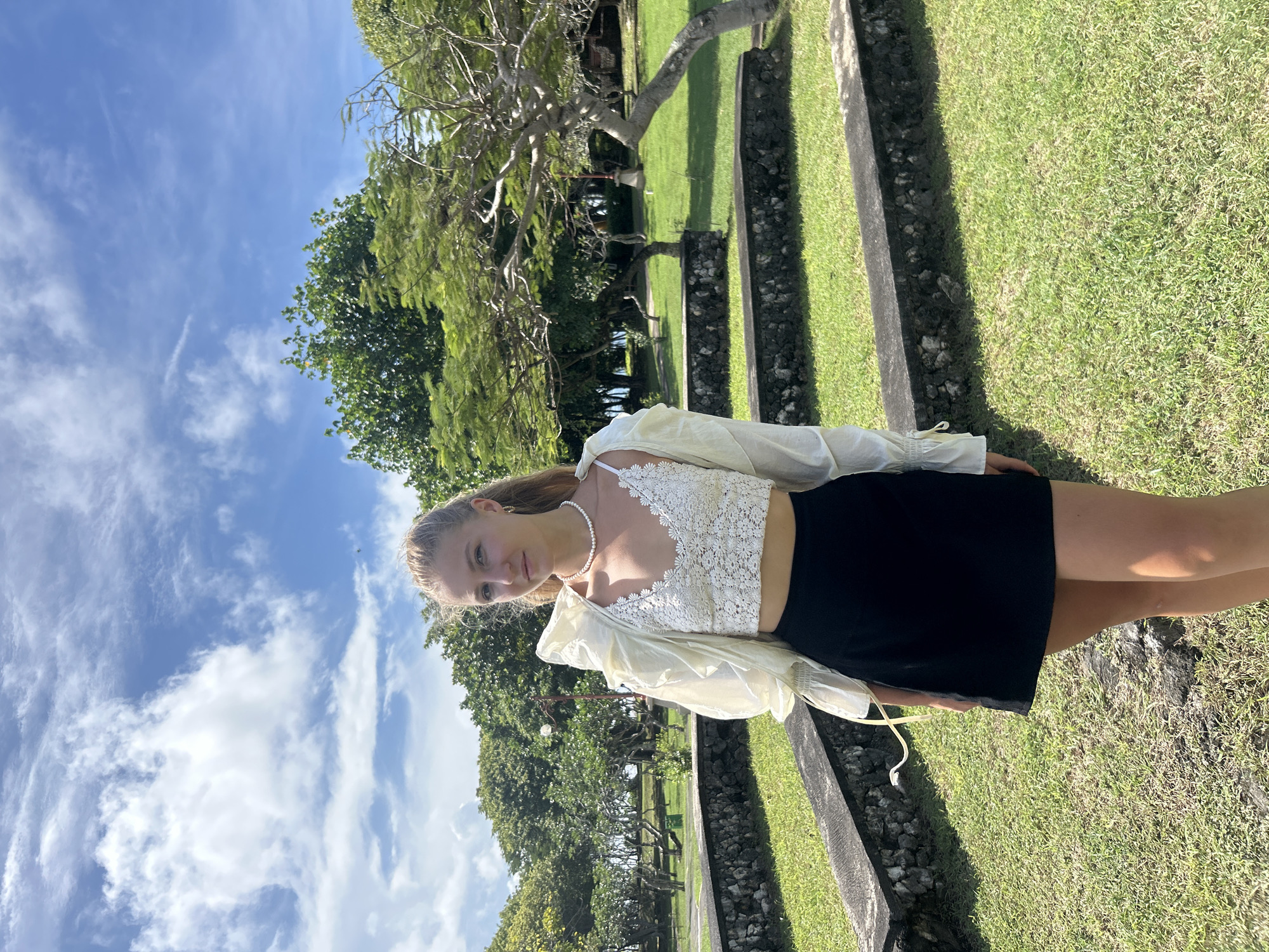 A young woman stands outdoors, posing for a photograph in what appears to be a park or well-maintained garden in Kuta Selatan, Indonesia. She is positioned slightly to the left of the frame, looking directly at the viewer with a gentle, neutral expression. She has blonde hair, pulled back, and is wearing a white lace crop top, a light-colored, possibly sheer, long-sleeved overshirt, and a black mini skirt. A pearl necklace is visible around her neck.
The background features a lush green lawn, neatly bordered by concrete or stone edges. Several trees, some with thick green foliage and others with more sparse, gnarled branches, are scattered throughout the grassy area. In the distance, beyond the trees, hints of water or a clearer horizon are visible. The upper portion of the image is dominated by a bright blue sky with scattered white, wispy clouds, indicating clear and sunny weather. Strong sunlight casts distinct shadows across the grass and along the concrete borders. No visible text is present in the image. The time of day is clearly daytime, likely mid-morning or afternoon, given the bright illumination.