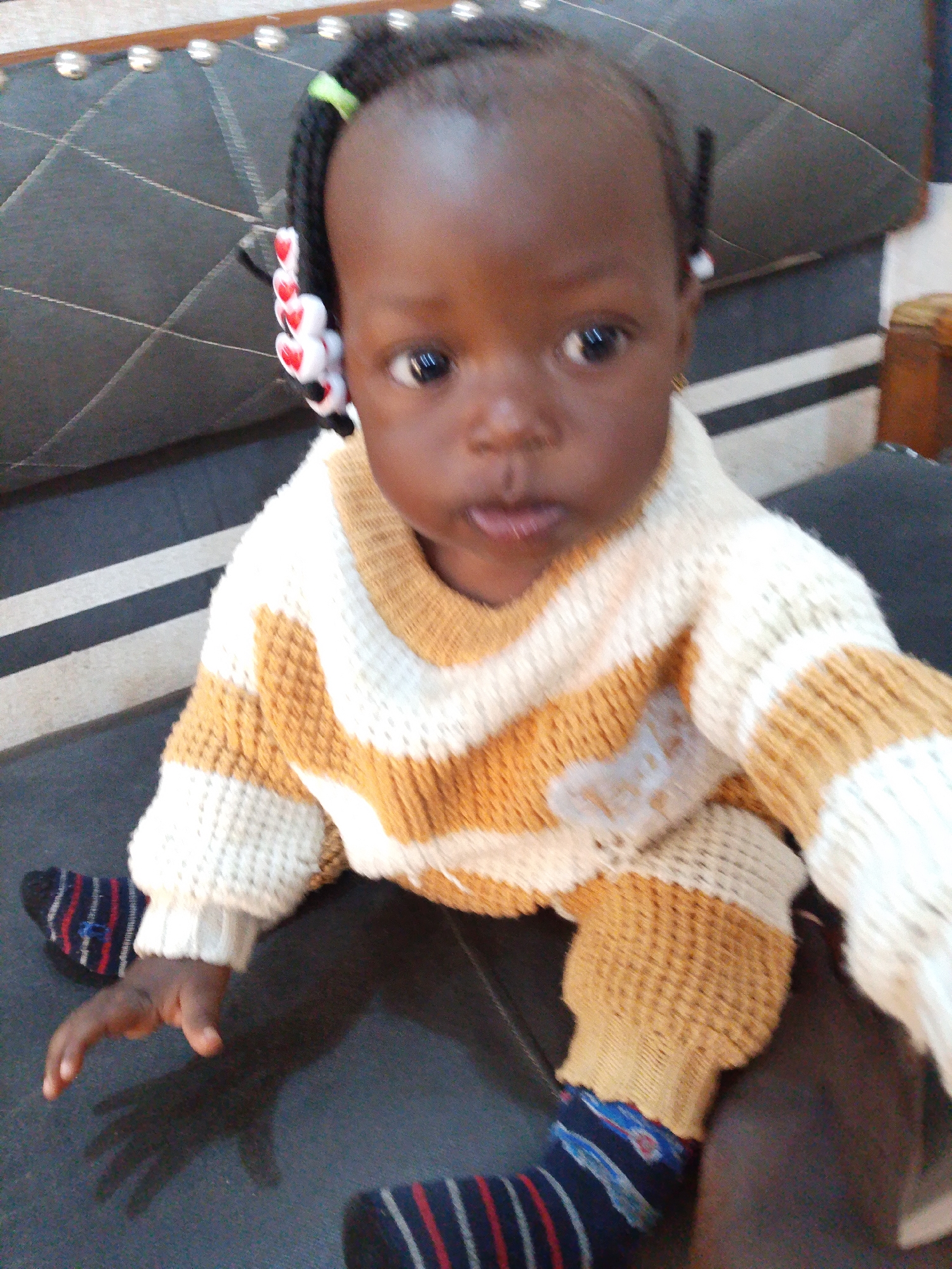 A close-up, eye-level shot shows a young Black child, likely a toddler, sitting on a dark, padded surface that appears to be a couch or chair. The child has dark skin and black hair styled into braids adorned with red heart-shaped beads and a green elastic band. The child is wearing a knitted sweater with horizontal stripes in off-white and a mustard yellow or light brown color. The child's legs are visible, and they are wearing socks with navy blue, red, and white stripes. The child's right hand is extended forward, casting a shadow on the surface. The background is out of focus but suggests an indoor setting with padded furniture and possibly a wooden cabinet visible on the right. The lighting is somewhat dim, suggesting indoor lighting. The image has a slightly grainy quality, consistent with a photograph taken with a mobile phone in low light. The specific location of Jos, Nigeria, is not directly evident from visual cues within the image itself; this information is provided as contextual data.