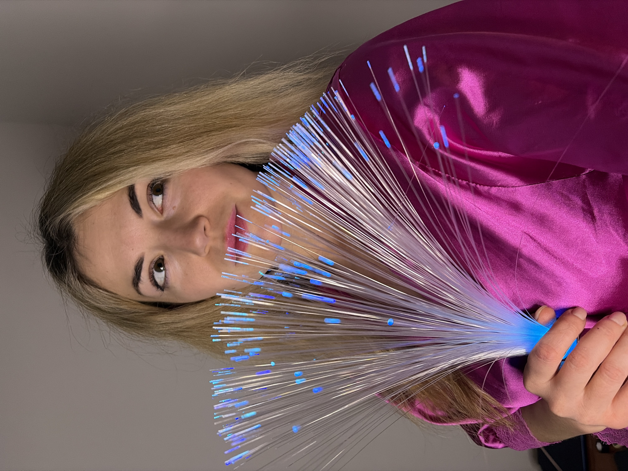 A fair-skinned woman with blond hair is holding a fiber optic light in front of her face. The light is emitting blue light from the tips of the fibers. She is wearing a pink, satin-like garment. The background is a plain, light gray wall. The location is not identifiable as Budva, Montenegro.