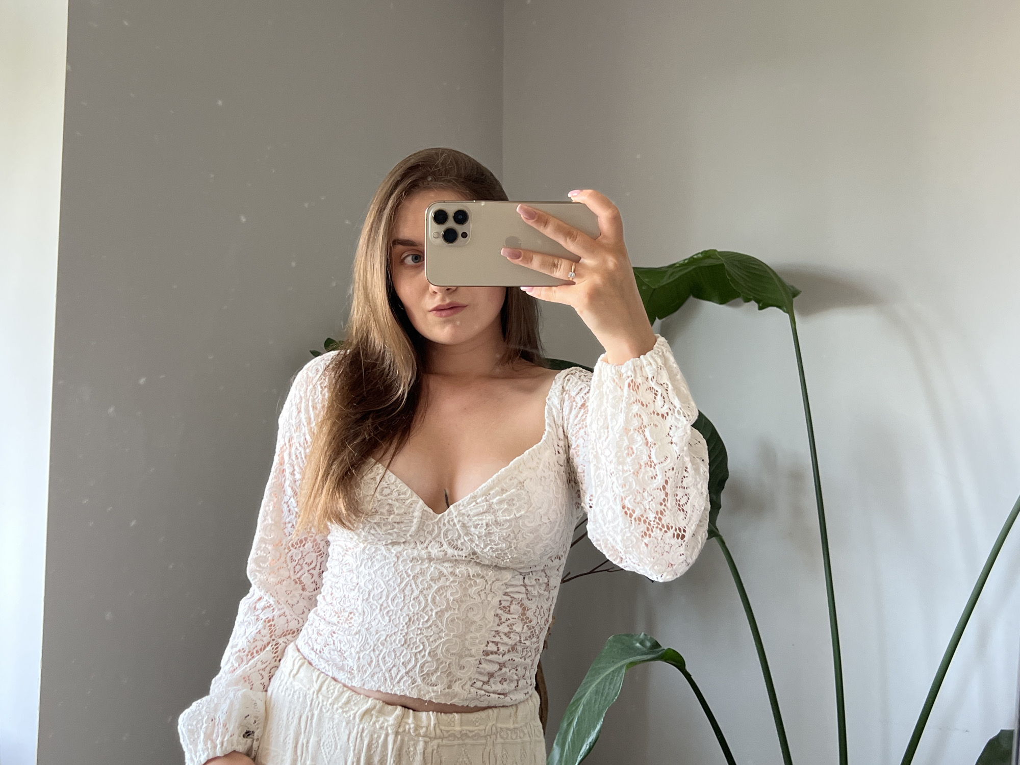 In Ternopil, Ukraine, a young woman with long, brown hair is taking a mirror selfie indoors. She is wearing a white lace top with voluminous sleeves and a v-neckline, and possibly cream-colored pants. She is holding a gold iPhone in her right hand, partially obscuring her face. Her left hand is visible, adorned with a ring on her ring finger and a light pink manicure. Behind her, a large green plant with broad leaves stands to the right, and the background is a plain grey wall with subtle speckles. The lighting suggests an indoor environment, possibly a room with natural light coming from the left.