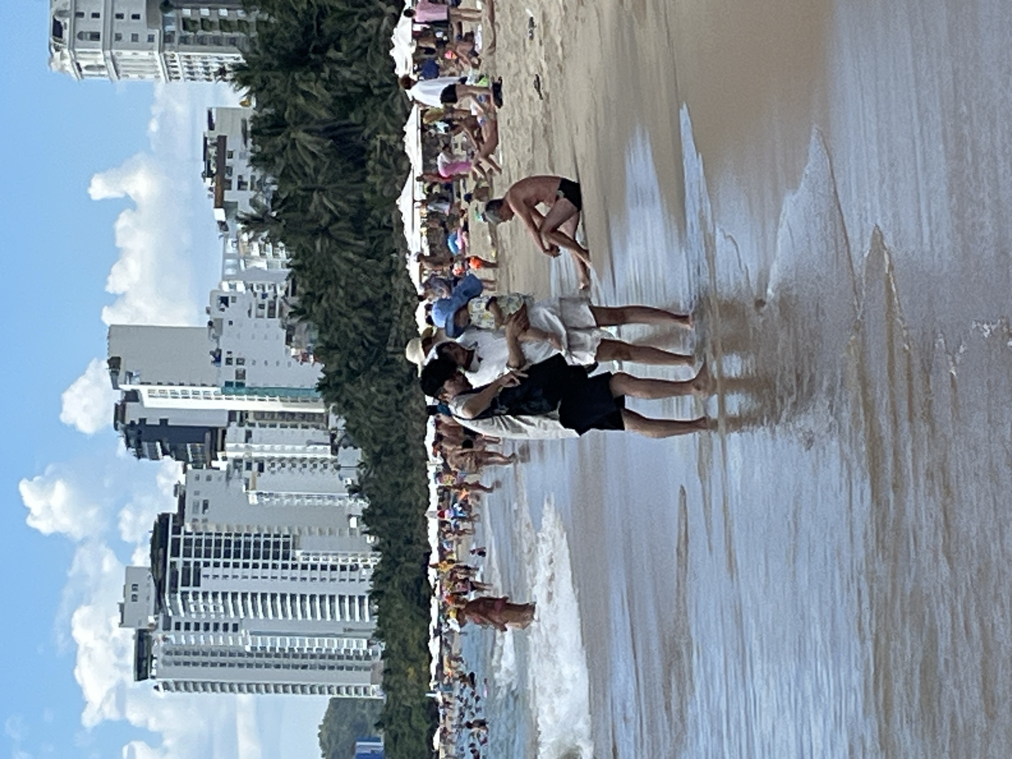 The image captures a sunny day at a crowded beach in Nha Trang, Vietnam, with modern high-rise buildings visible in the background, characteristic of the city's coastal skyline. In the foreground, a family of three stands near the water's edge. A man in a white shirt and black shorts holds a young child wearing a blue hat. Beside them, a woman in a white top and white shorts appears to be interacting with the child. A man in black shorts is seated on the sand further back, facing away from the water. The beach is populated with many people swimming, sunbathing, and walking, with white umbrellas dotting the sand. The ocean exhibits gentle waves rolling onto the shore. The overall scene depicts a lively beach day in a vibrant urban coastal environment.