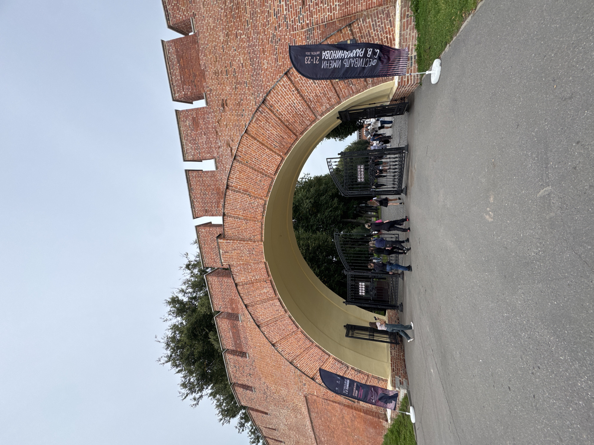 The image shows an arched gateway made of red brick, which appears to be part of a historical fortification. The gateway is open, revealing a black metal fence and people walking through. There are two large flags on stands outside the gateway, one on each side. The flag on the right has text that reads "ФЕСТИВАЛЬ ИМЕНИ 21-23 С. В. РАХМАНИНОВА АВГУСТА 2026," indicating it is a festival in honor of S.V. Rachmaninoff held on August 21-23, 2026. The gateway is located in Veliky Novgorod, Russia. The sky is overcast, and there are trees visible behind the gateway. A few people are walking through the gateway and on the pavement in front of it. One person on the left is holding a camera. The ground is paved with asphalt.