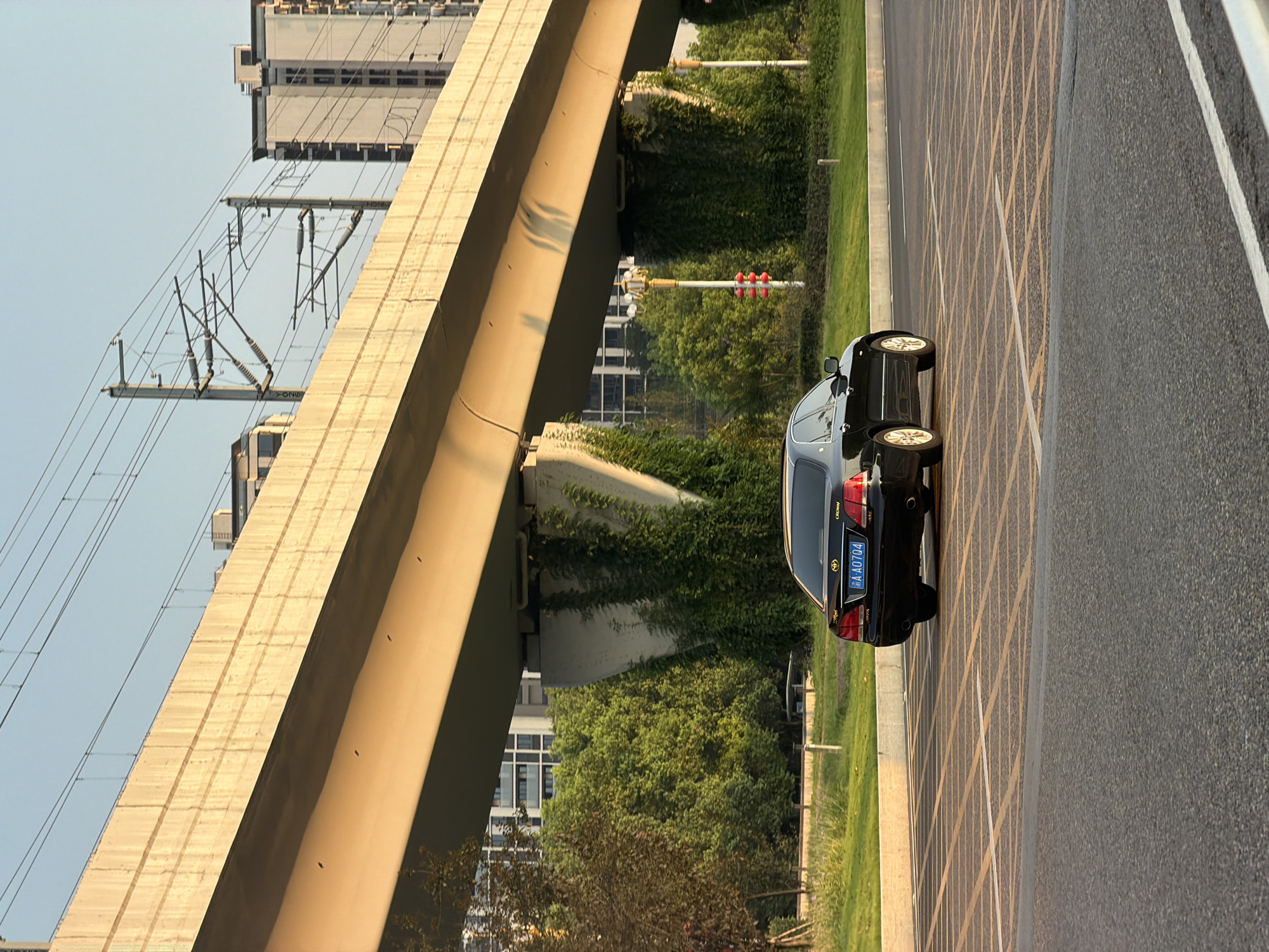 A black Toyota Crown sedan is parked on a street with yellow and white striped parking lines in the foreground. The car's license plate reads "AA0704". Behind the car, a large concrete overpass runs horizontally across the frame, casting a shadow on the road. The overpass supports train tracks and power lines. The supports of the overpass are covered in vines. In the background, there are several tall, modern buildings and lush green trees. The scene is illuminated by the warm light of what appears to be a sunset or sunrise. The environment suggests an urban setting in Jiangjin, China, likely near a transportation hub. There are no people visible in the image.