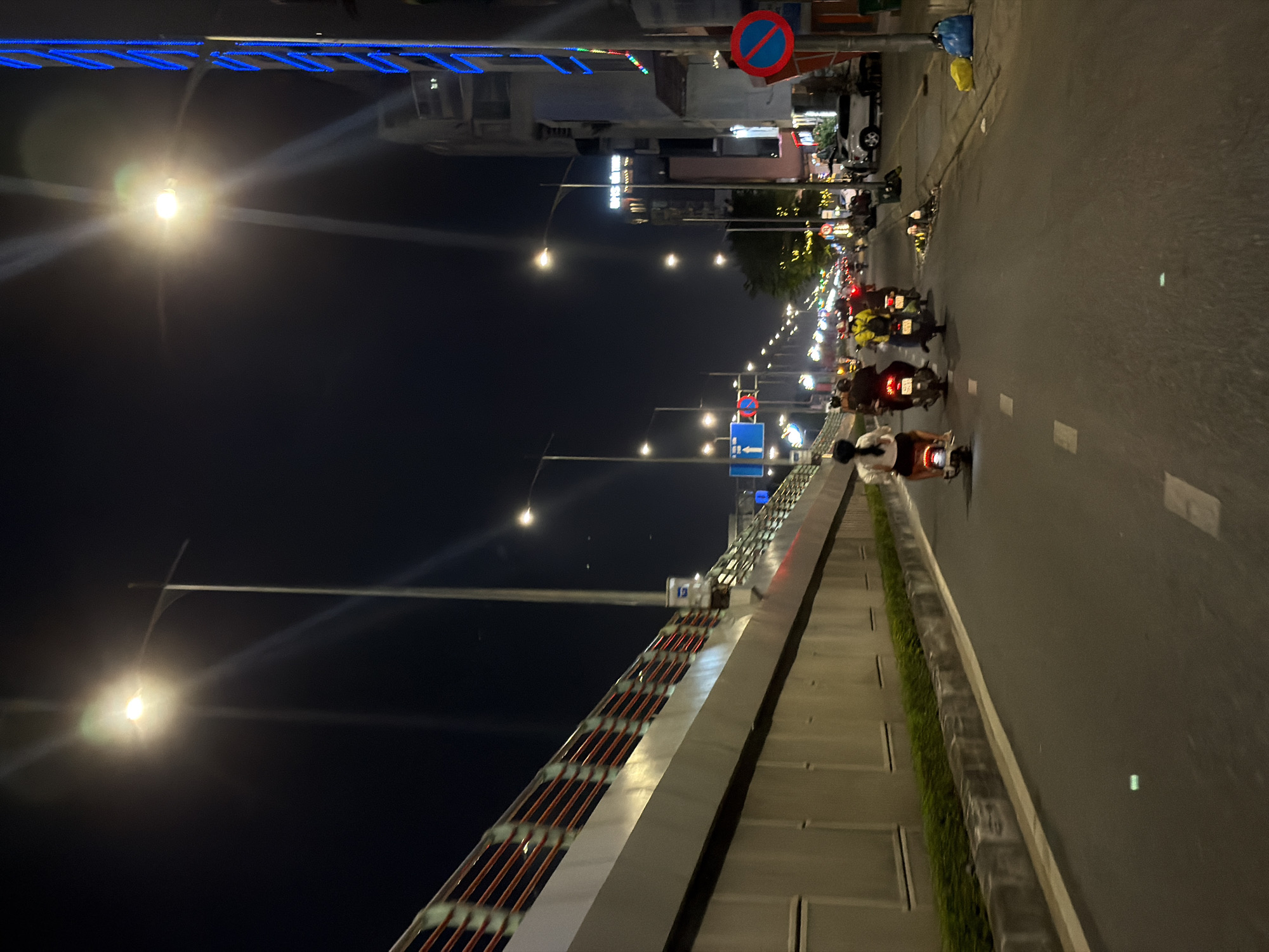 This media file captures a nighttime scene on a busy street in Da Nang, Vietnam. The image is oriented vertically, with the street and its activity filling the right side, and a bridge or elevated walkway occupying the left.
The primary subjects are numerous motorbikes navigating the road. Many of the riders are visible, some wearing helmets. The motorbikes' headlights and taillights create streaks of light, indicating movement and the low-light conditions. A few cars are also present in the background. The street is lined with lampposts, casting a warm glow and illuminating the scene. Along the left side of the frame, a bridge structure with red railings and metal supports is prominent, with blue decorative lights running along its top edge. Some buildings are visible in the distance beyond the street.
The activity is the flow of traffic on a city street at night. The time of day is clearly night due to the artificial lighting and the dark sky. The weather appears clear, as there are no indications of rain or fog.
Notable details include the traffic signs visible, such as a red circle with a blue interior and a red diagonal line, indicating a prohibition, and blue directional signs. The presence of many motorbikes suggests a common mode of transportation in this urban environment. The decorative blue lights on the bridge add a modern aesthetic to the city's infrastructure. The overall mood is one of a vibrant, active urban nighttime environment. No legible text is clearly discernible beyond the traffic signs.