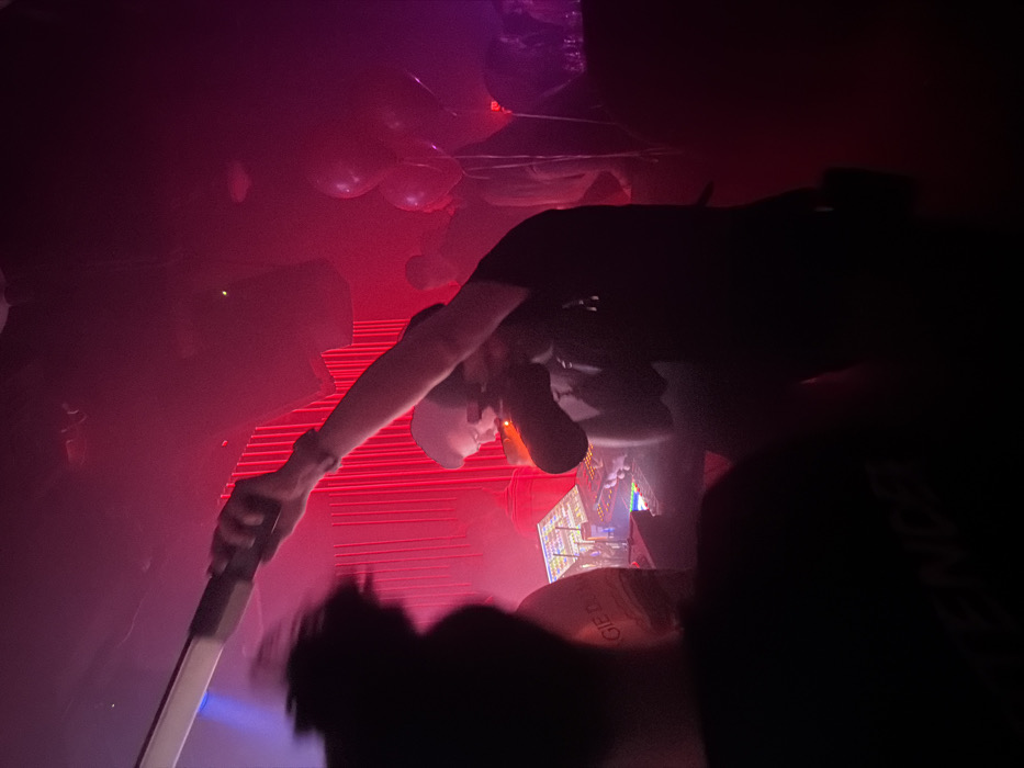 The image depicts a crowded, dimly lit indoor scene within an entertainment venue or nightclub in Moscow, Russia. Red and purple ambient lighting predominates, with some blue light visible in the upper left. A haze or smoke is present in the air.
In the center, a male individual, appearing to be a DJ, stands behind a multi-screen DJ console displaying various colored interfaces. He wears a dark shirt and has dark hair. His left hand holds a dark cylindrical object, possibly a microphone or drink, to his mouth, while his right arm is extended upwards, holding a white rectangular light baton or similar elongated device.
To the left, a person with dark, curly hair is seen from behind, facing towards the DJ console. The back of their dark shirt shows partially visible white text at the bottom.
In the background to the right, several dark balloons are visible near the ceiling. Indistinct silhouettes of other individuals are present throughout the background, suggesting a crowd. Large dark speakers or light fixtures are mounted high on the walls. The wall behind the DJ features vertical red lighting elements, possibly curtains or light strips. A white object on the DJ console bears visible text that reads "GIE DUM" and "Moscow".