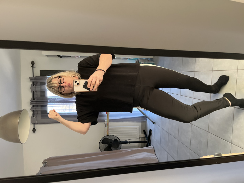 The image displays a reflection of a person taking a selfie in a mirror, which is oriented horizontally. The person, who has short blonde hair and is wearing dark-rimmed glasses, is positioned centrally within the mirror's frame. They are wearing a black short-sleeved top or sweatshirt, black leggings with a thin white vertical stripe down the outer side of each leg, and black socks. A black beaded bracelet is visible on their right wrist.
The individual is holding a light-colored smartphone with a dark case in their right hand, using it to take the picture. Their left arm is bent at the elbow, with the fist clenched, appearing as if flexing a muscle. The person's face shows a playful expression, with their lips pursed in a "fish face" or pout.
The background, visible through the mirror's reflection, reveals a room with light gray tiled flooring. On the left side of the reflection, a metallic, dome-shaped lamp shade is prominent. A window is visible in the middle ground, covered by dark gray horizontal blinds. Below the window, a light-colored radiator or heater can be seen. To the right of the person, a black standing fan is visible on the floor. Further into the background, what appears to be a bed or sofa with a gray throw is partially visible. No discernible text is visible in the image. The lighting suggests it is daytime.