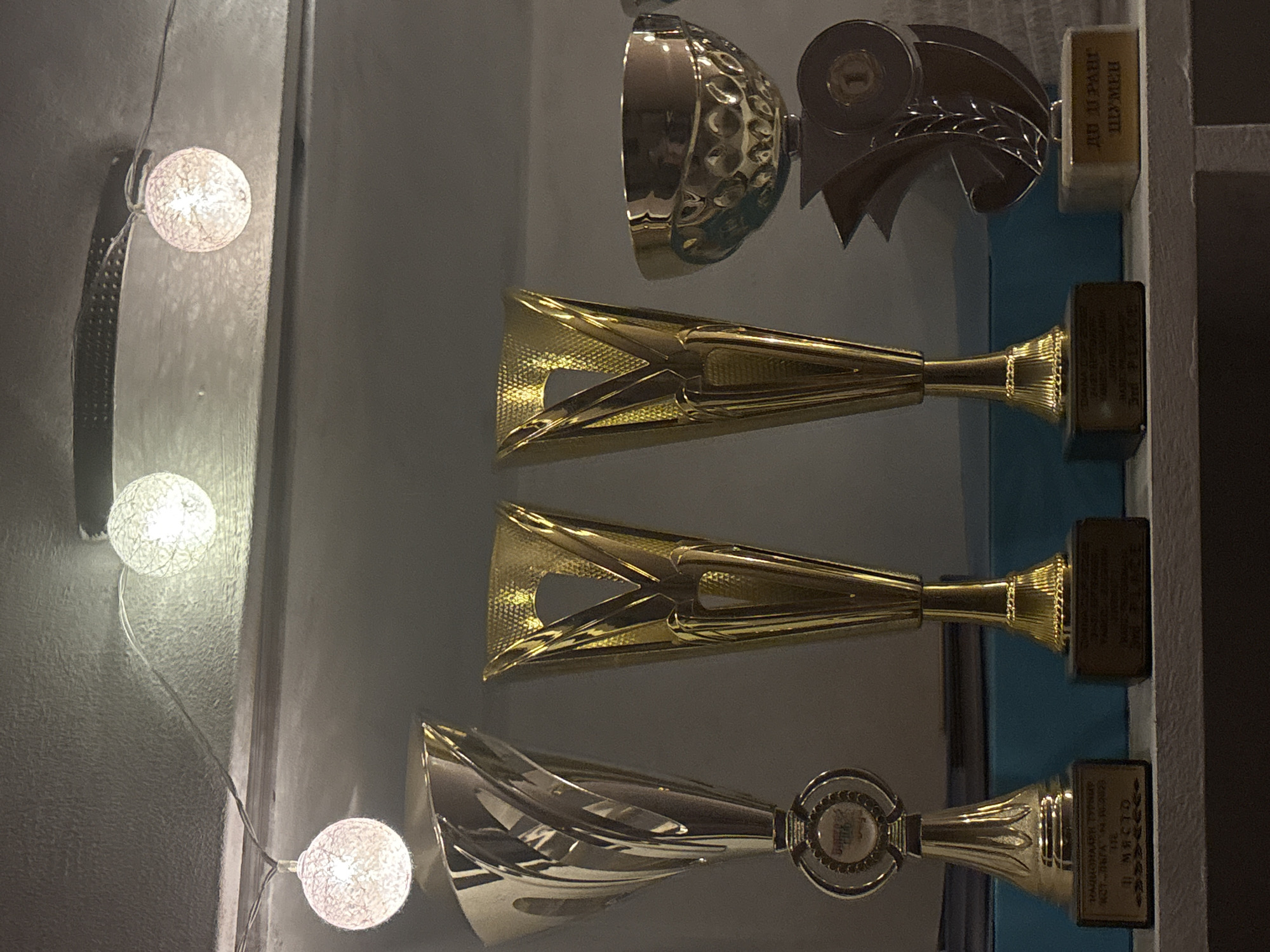 An indoor, eye-level, medium shot captures a shelf displaying four trophies and a string of globe-shaped lights overhead. The trophies, from left to right, include a silver trophy with a flared top, a tall gold trophy with a V-shaped opening, another similar tall gold trophy, and a smaller gold cup with decorative indentations. The trophy bases have inscriptions, though they are partially obscured. The background is dark and indistinct. The lighting is dim, with the globe lights providing a soft glow. The trophies are arranged on a white shelf, with blue folders visible behind them. The scene appears to be in a domestic or informal setting.