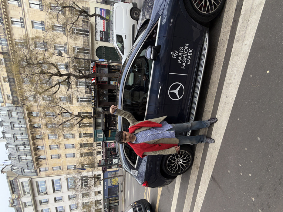 An urban street scene in Paris, France, is captured, with a prominent view of the Théâtre des Variétés located at 7 Boulevard Montmartre. A young adult male stands on a white-striped pedestrian crossing, positioned left of a dark blue Mercedes-Benz EQE SUV.
The male subject, possessing dark hair and a neutral to serious facial expression, is attired in a light beige blazer over a blue and white striped button-down shirt, with a bright red knit sweater draped over his shoulders. He wears blue denim jeans and dark, studded slip-on shoes. His right arm is bent, with his hand raised and fingers pointing downwards towards the ground or the vehicle.
The Mercedes-Benz EQE SUV features a prominent white Mercedes-Benz star logo on its side, accompanied by the text "FW PARIS FASHION WEEK," indicating its official association. A silver running board is visible along the lower edge of the vehicle's side. A reflection of a person is visible in the driver's side window.
The background consists of multi-story Haussmannian buildings with light-colored stone facades, numerous windows, and grey mansard roofs. Storefronts are visible at street level, including a clear sign for "THÉÂTRE DES VARIÉTÉS" and a partial sign reading "PANORAMA." Deciduous trees with bare branches line the street. A white utility van is visible further down the road, and another vehicle is partially visible on the far left. The sky is overcast.