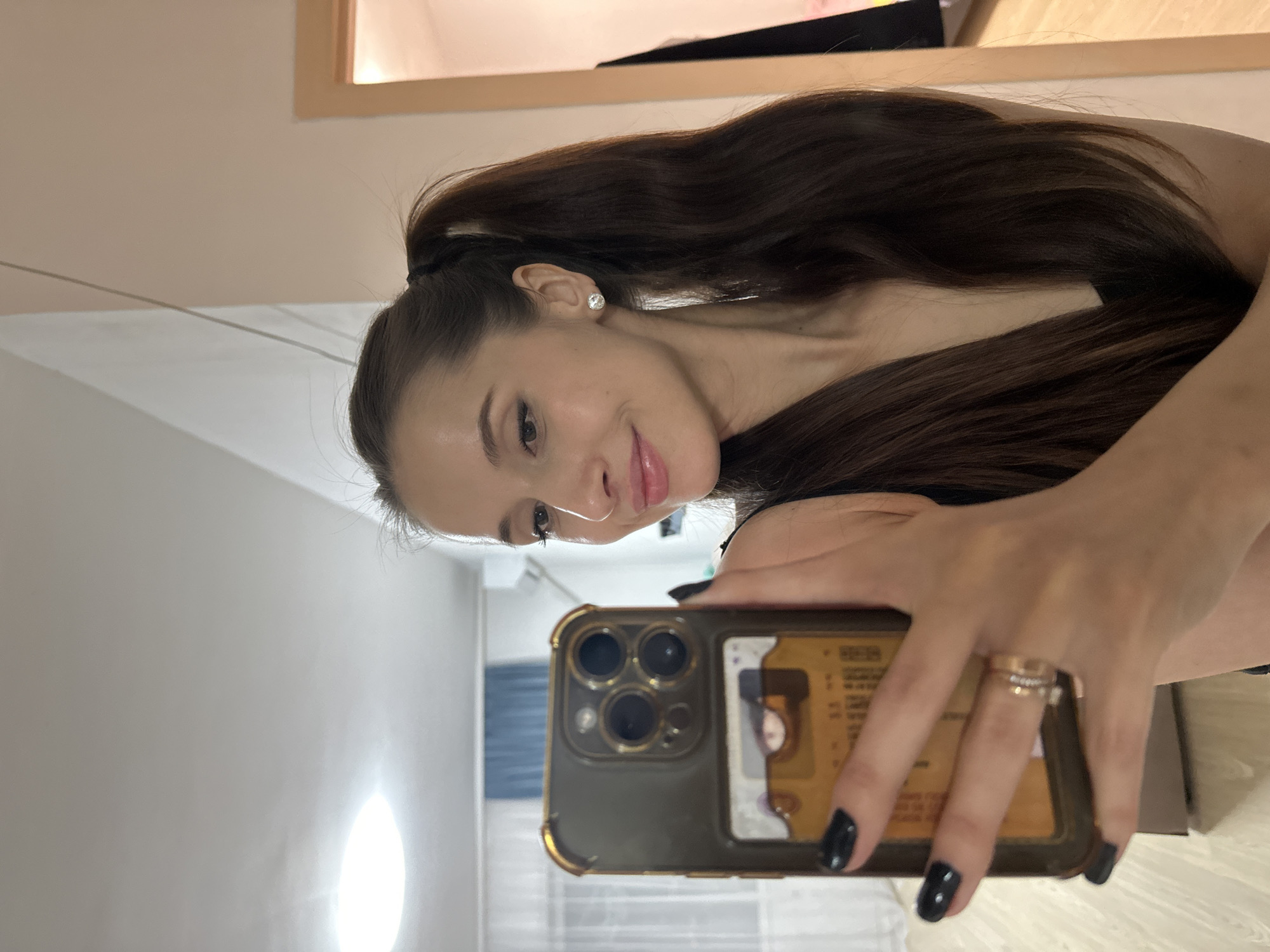 A woman with long brown hair pulled back into a ponytail is taking a selfie with an iPhone. She is wearing a black top and has a ring on her finger. The phone has a brown case and a card inserted in the back. The background appears to be a room with a white wall, a window with blinds, and a cabinet. The location is likely indoors. There is no information to determine if the photograph was taken in Yekaterinburg, Russia.