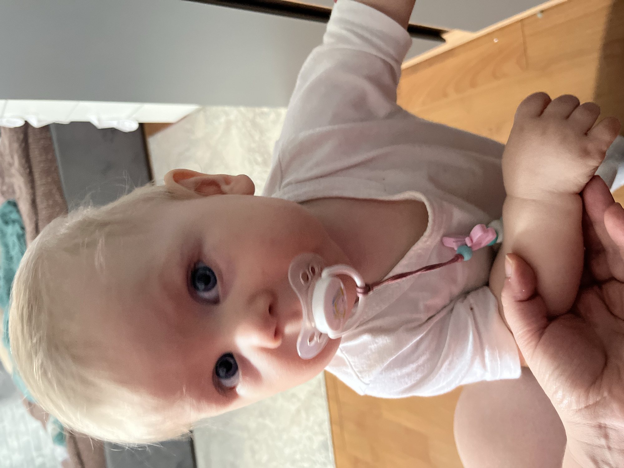 A fair-skinned baby with blonde hair and blue eyes is looking at the camera with a pacifier in their mouth. The baby is wearing a white shirt. An adult hand is visible in the lower right corner, holding the baby's hand. The background includes light-colored wooden flooring and a partially visible white cabinet or wall. The scene is indoors. The location is unknown; no information in the image supports it being in Vesyoloye, Russia.