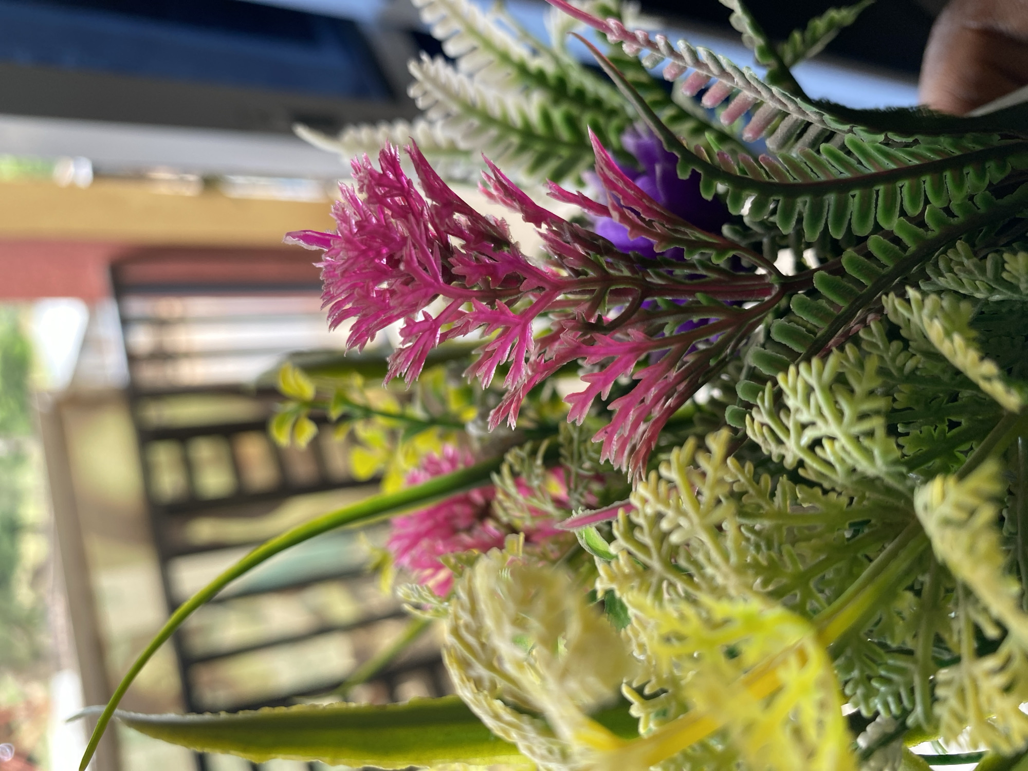 The image is a close-up of artificial flowers, including pink, purple, yellow, and white foliage. The background is out of focus and shows a building exterior with window frames. Given the prompt's request to include a location in Bukuru, Nigeria, but with no indication that the image comes from there, it is impossible to confirm if this photo was taken there. Thus, I will refrain from stating that it was.