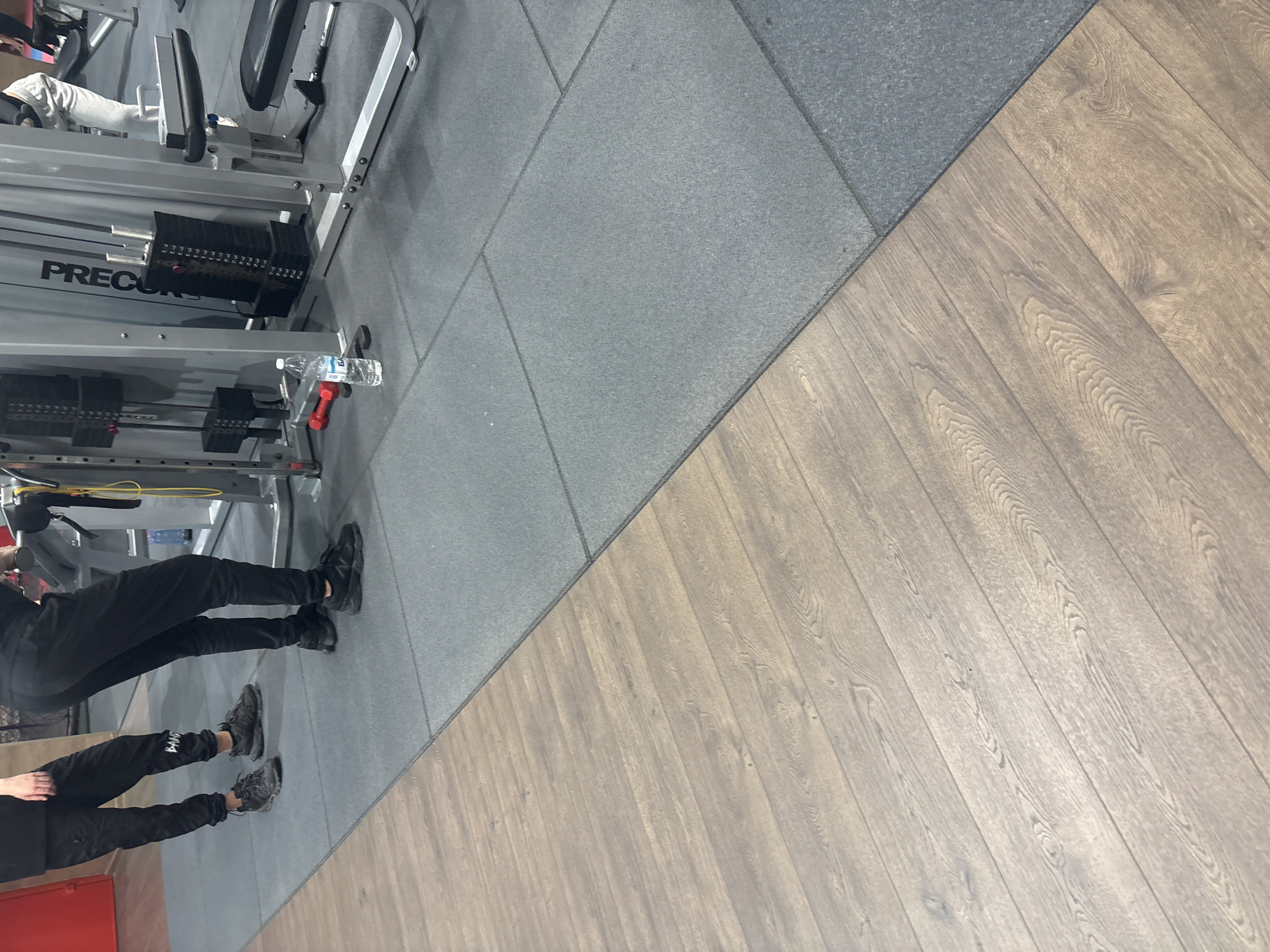 The image shows a section of a gym, likely indoors. A "Precor" branded weight machine is visible, with weight plates stacked. Two people are present, wearing dark athletic clothing and shoes. One person appears to be using the weight machine, while the second is standing nearby. The floor is divided into two sections, one with gray rubber matting and the other with wood-patterned flooring. The presence of the weight machine and athletic clothing suggests a fitness environment. There is no indication of the city of Varna, Bulgaria, within the image.