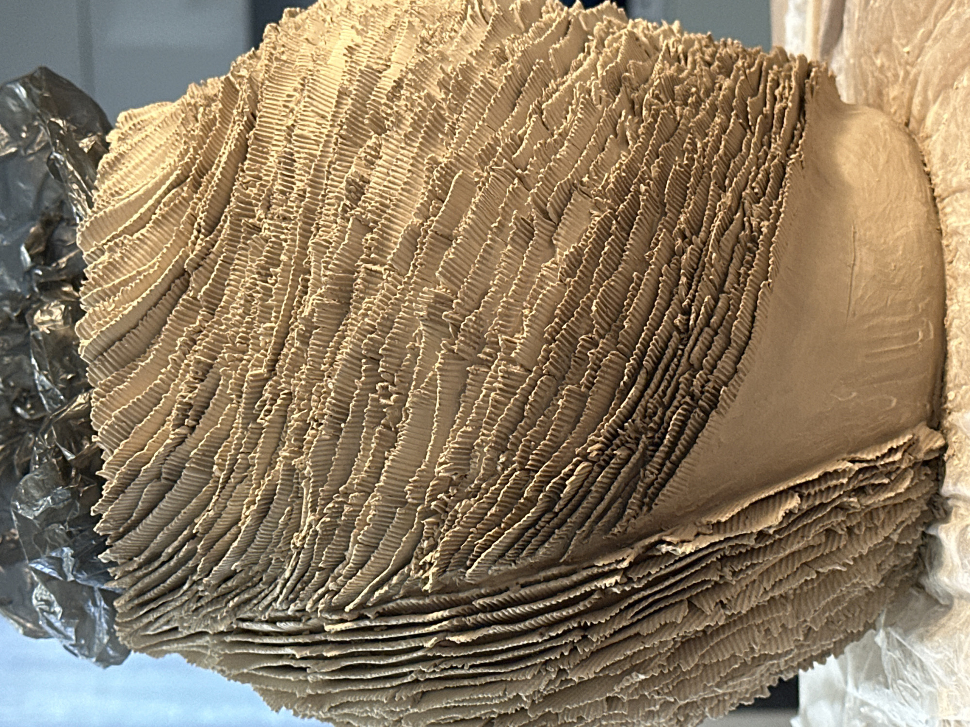 The image shows a close-up of a textured, light brown, rounded object, possibly a sculpture or a craft project. The surface is covered in numerous overlapping, corrugated layers that create a ribbed effect. The object has a rough, unglazed texture, and appears to be made of clay or a similar material. At the top, there is a crumpled dark gray material, which could be foil or plastic wrap, possibly covering part of the object. The object is resting on a white surface, which appears to be paper or fabric, in an indoor setting. The background is out of focus, with hints of blue and gray, suggesting it could be a wall or window. There is no indication of the city of Nevinnomyssk, Russia, in the image.