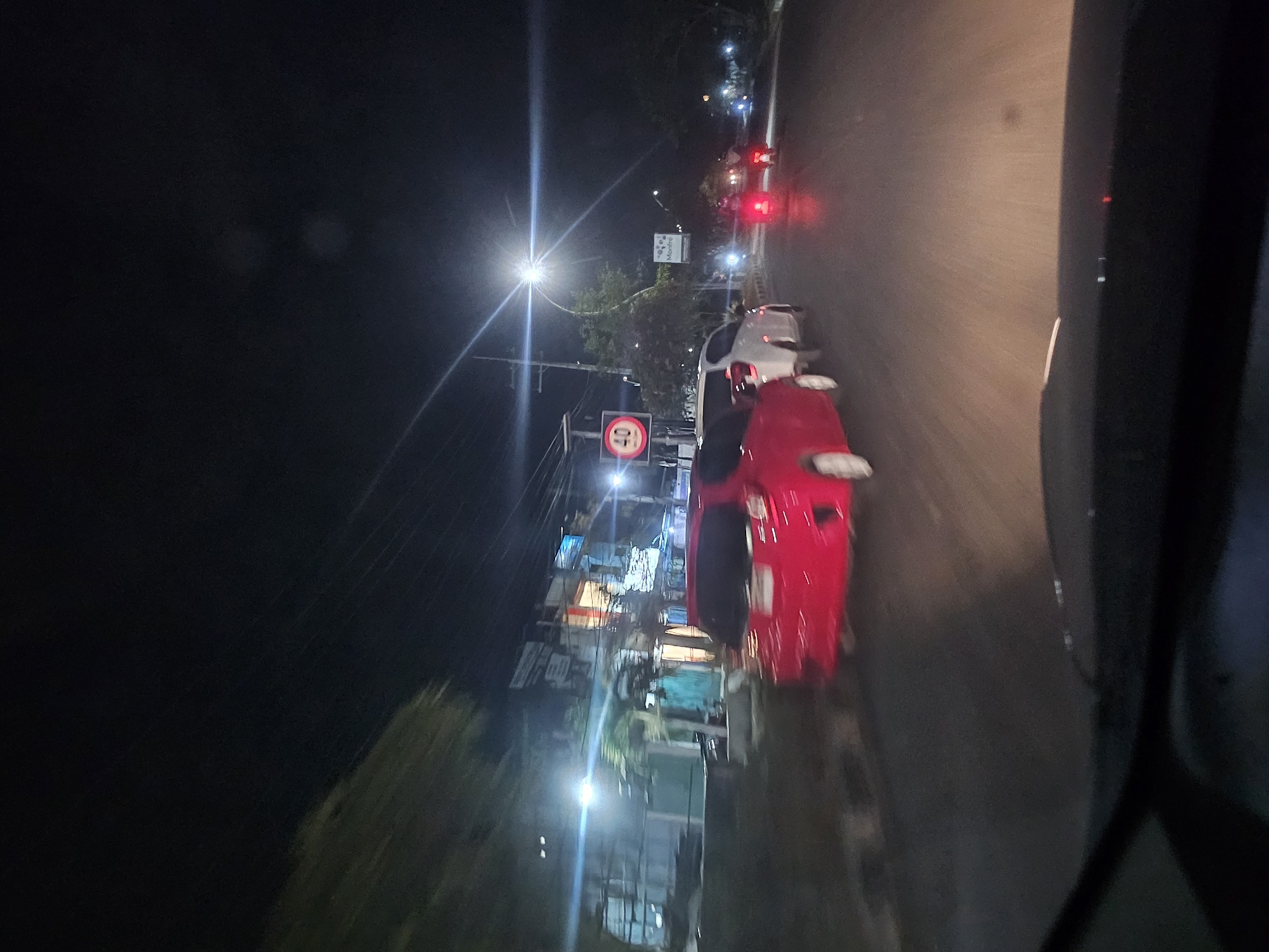 The image depicts a night scene on a road in Sukomulyo, Indonesia, likely captured from inside a vehicle. The foreground is dominated by the dashboard of a car, suggesting the photographer is a driver or passenger. On the road, there are several vehicles. A red hatchback is prominently positioned in the left lane, with a white car following closely behind it. Further down the road, two motorcycles are visible, their headlights and taillights illuminating the asphalt. The environment appears to be a moderately built-up area with buildings lining the left side of the road. Streetlights are present, casting a glow on the surroundings. A speed limit sign indicating "40" is visible on a pole to the left. The overall impression is that of a typical urban night drive in Indonesia.