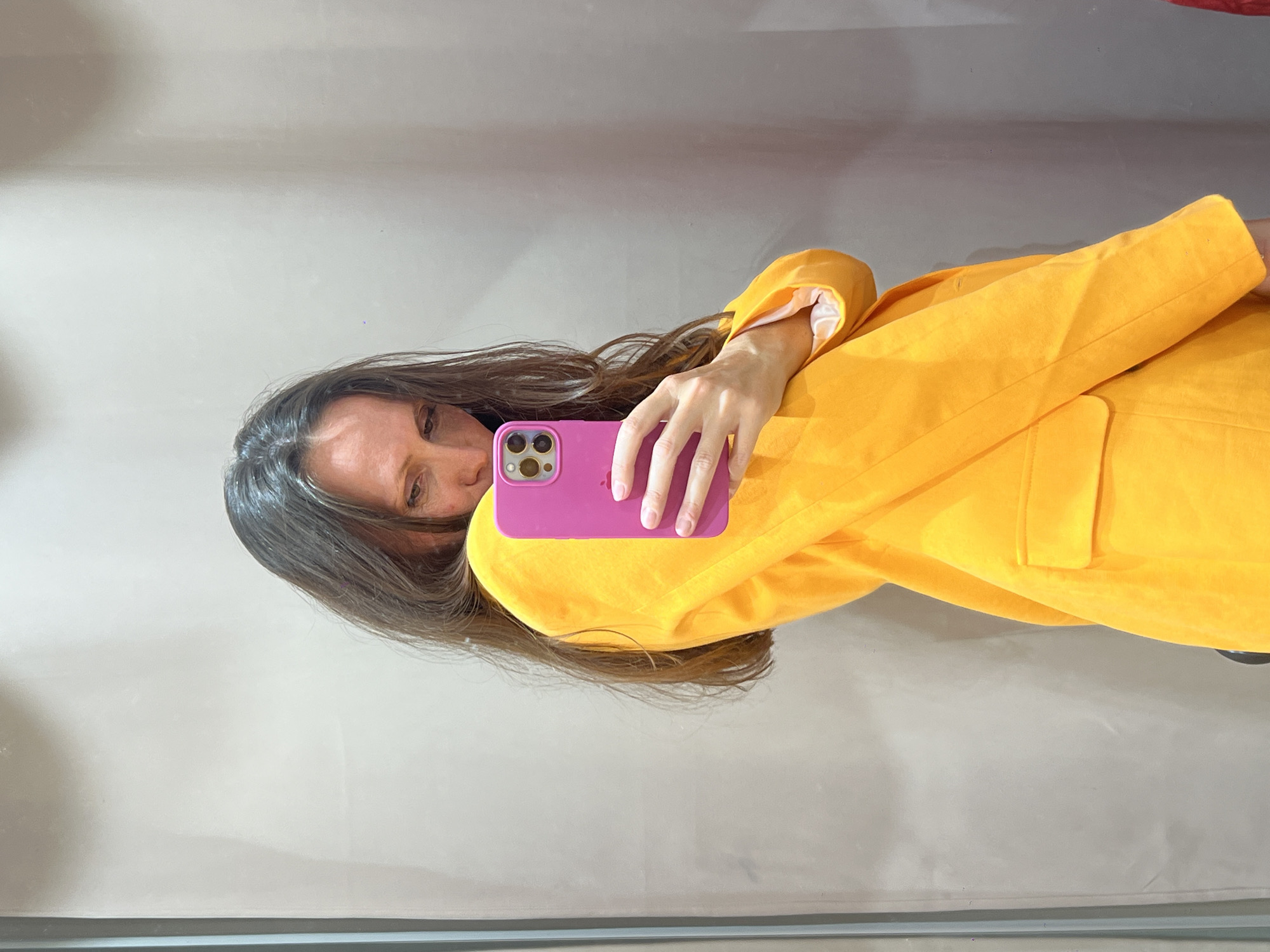A woman is taking a selfie in front of a mirror. She is wearing a bright yellow blazer. She is holding a pink iPhone. The background is a plain, light gray wall. The location cannot be determined from the image.