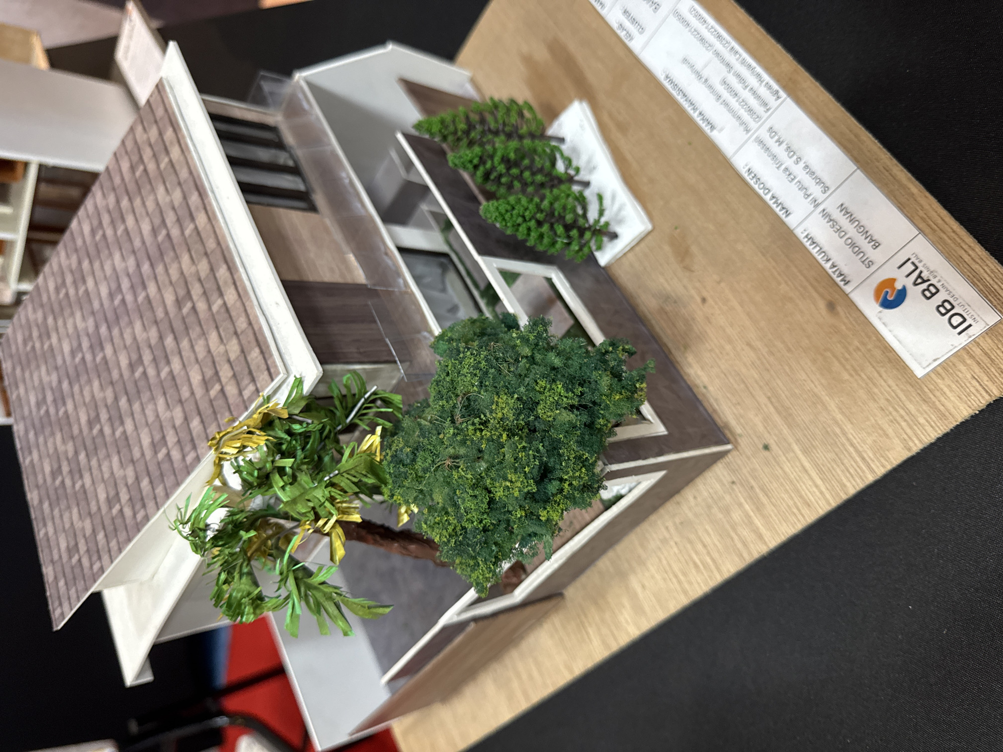 The image shows an architectural model sitting on a light brown wooden surface, which transitions into a black surface on the right. The model appears to be a contemporary house design, featuring a brown tiled roof, white trim, and several sections with varying heights and angles. There are miniature trees included in the model's landscaping, giving the impression of a well-planned outdoor space.
A white label is placed beside the model, bearing the logo of "IDB BALI" (Institut Desain & Bisnis Bali), indicating that the model is likely associated with the design and business institute in Bali. Text on the label includes sections labeled "MATA KULIAH" (subject), "NAMA DOSEN" (lecturer name), and "NAMA MAHASISWA" (student name), with the names "Ni Putu Eka Trisnasari," "Subrata, S.Ds, M.Ds," "Fallistea Fidian Santosa," and "Agnes Hariyanti Laila" followed by numbers in parentheses, potentially student ID numbers.
Given the location in the "IDB BALI" label, it can be inferred that the model is related to design studies or projects in Denpasar, Indonesia, where the institute is located. There are no people visible in the image.