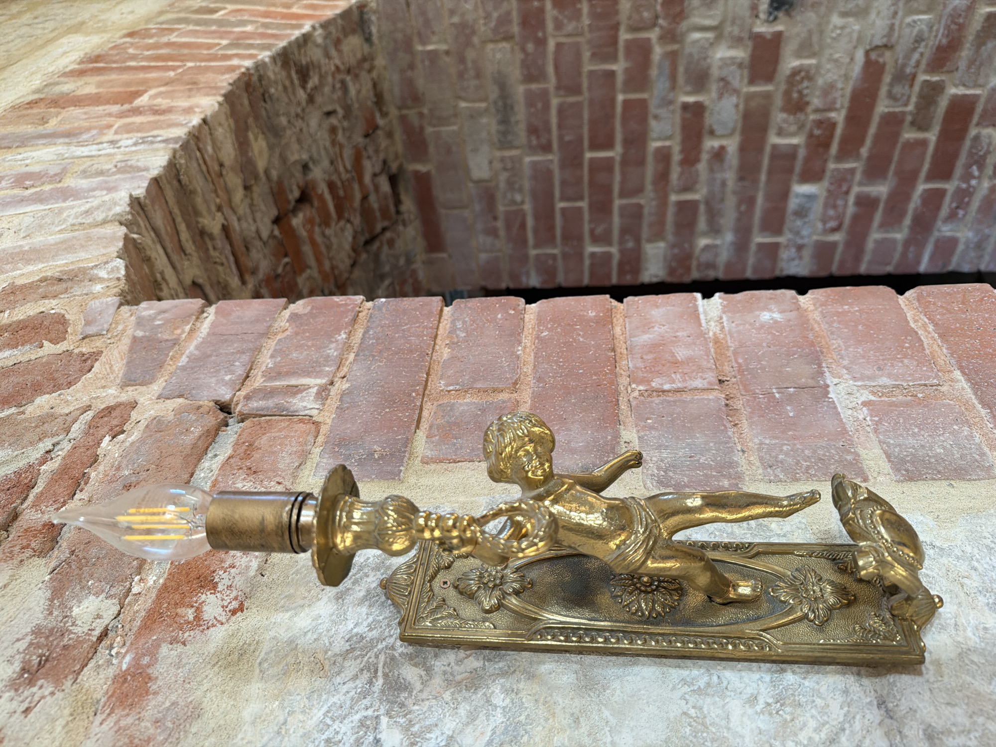 An antique brass wall sconce featuring a cherub figure is mounted on a textured brick wall in the village of Skoryakovo, Russia. The sconce has a decorative backplate with floral motifs and a cherub appearing to hold onto a decorative chain. A filament light bulb is affixed to the top of the sconce. To the right of the sconce, an arched brick opening reveals a darker interior with more brickwork. The overall scene suggests an old, possibly historical, building.
