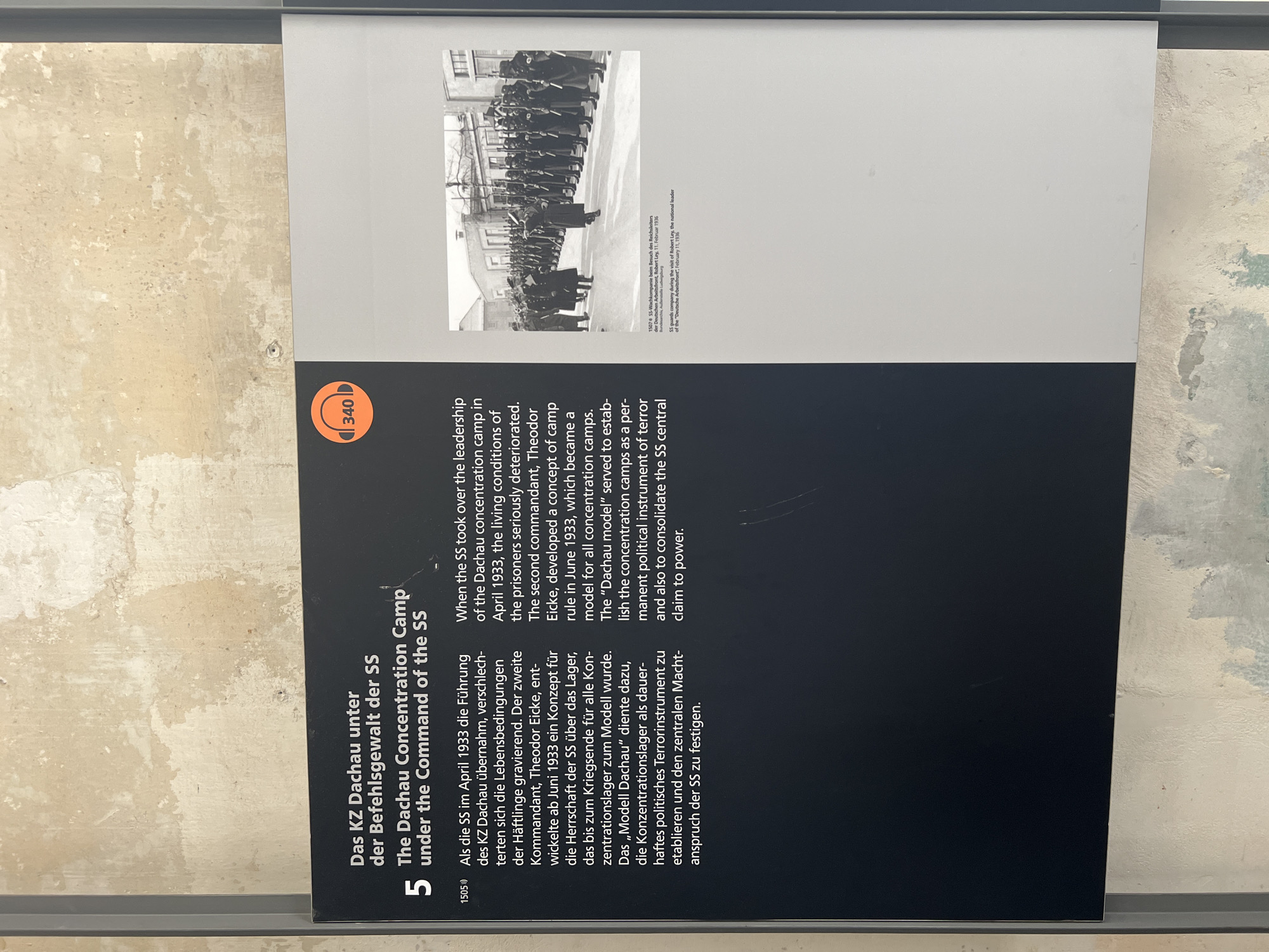 The image shows a panel from a museum exhibit about the Dachau Concentration Camp. The panel features text in both German and English, detailing the camp's operation under the SS. The heading reads "Das KZ Dachau unter der Befehlsgewalt der SS" and "The Dachau Concentration Camp under the Command of the SS." A large number '5' is also prominently displayed. Below the heading, two paragraphs of text describe the SS taking over leadership in April 1933, the deteriorating conditions, and the development of the "Dachau model" as a tool for political terror.
To the right of the text, there is a black and white photograph. The photograph depicts a group of SS guards in uniform, standing in formation with rifles, saluting. In the foreground, several SS officers are visible, with one appearing to be inspecting the troops. A building with a barred window is visible in the background.
Below the photograph, there is a caption in German and English that identifies the image as "SS-Wachkompanie beim Besuch des Reichsleiters der Deutschen Arbeitsfront, Robert Ley, 11. Februar 1936" and "SS guards company during the visit of Robert Ley, the national leader of the 'Deutsche Arbeitsfront,' February 11, 1936."
An orange circular icon with the number '340' in white is visible in the upper right corner of the text area, suggesting it might be an audio guide marker or exhibit number. The background of the exhibit panel appears to be a textured wall, possibly depicting aging plaster or stone. The exhibit is likely located in Dachau, Germany, given the subject matter.