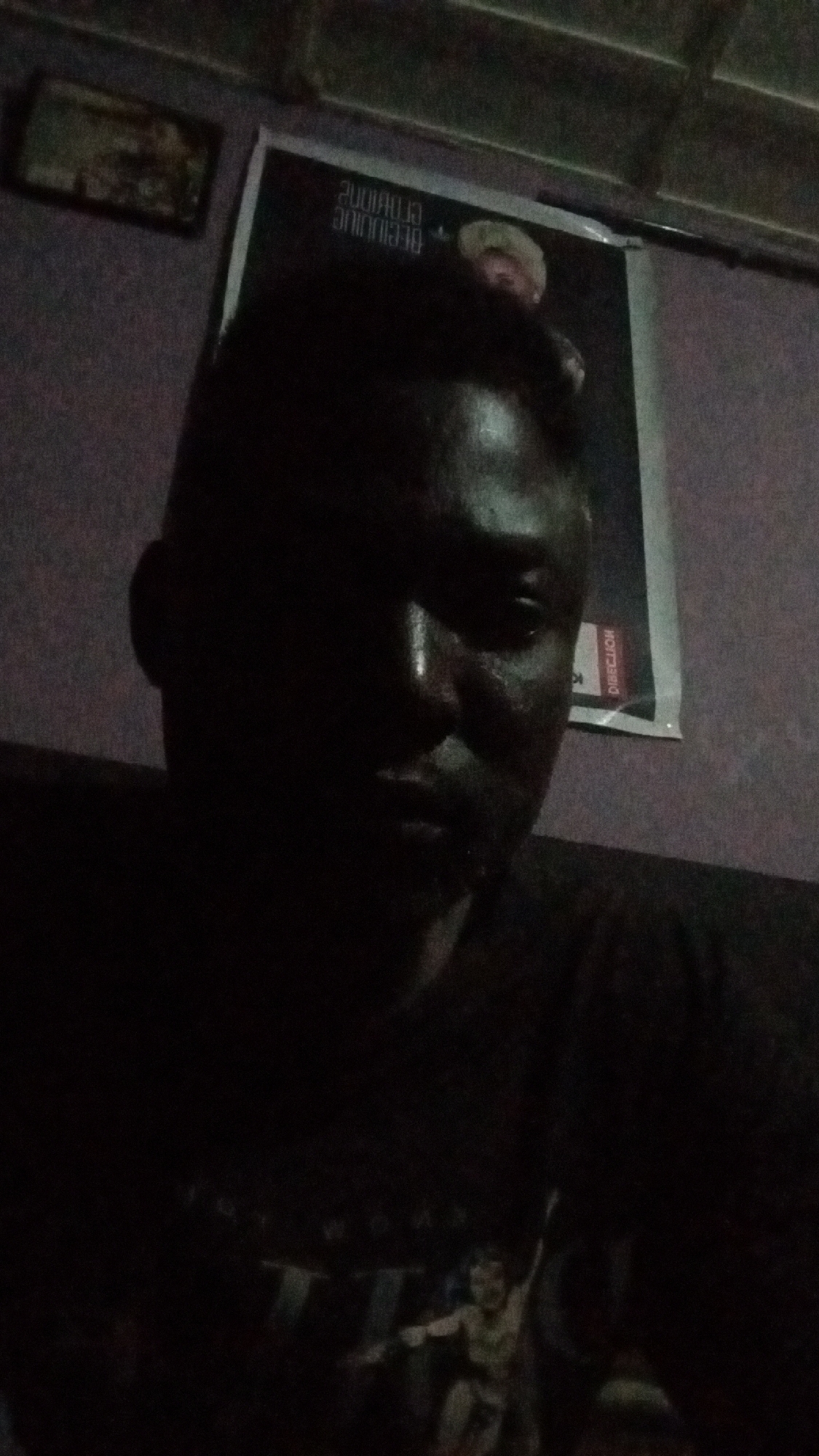 A dimly lit indoor scene features a close-up of a dark-skinned man. He is wearing a t-shirt with a graphic design of a person. Behind him is a poster on the wall with writing in reverse. Further back, above the poster, is a framed picture. The lighting is poor, making it difficult to discern details. The location is Sabon Tasha, Nigeria.