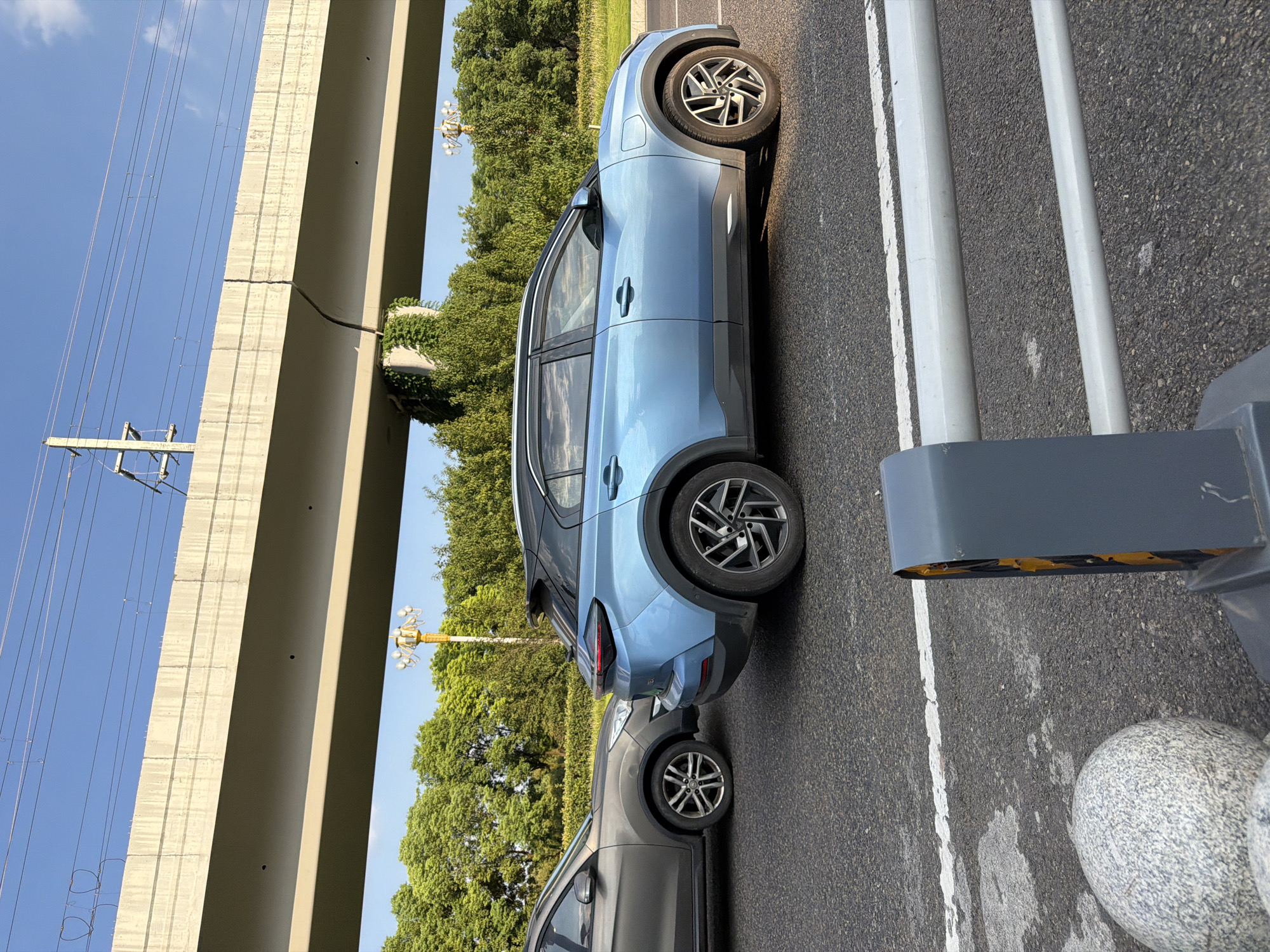 A blue electric SUV and a gray SUV are parked on a city road in Jiangjin, China, beneath an elevated concrete structure. The sky is blue with scattered clouds. Trees line the background, and a lamppost is visible. In the foreground, a concrete barrier with a traffic signal is present. The image appears to be taken during daylight hours.