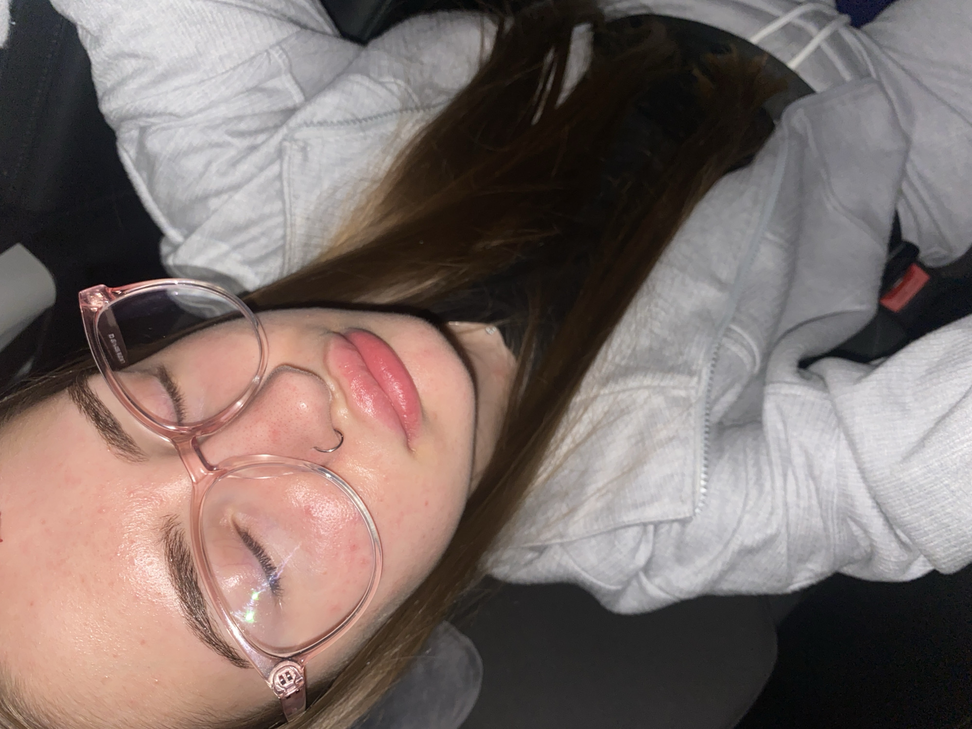 The image shows a close-up, top-down view of a young woman with her eyes closed, wearing large, pink-rimmed glasses. She has on a light gray hooded sweatshirt. Her hair is long and brown. A small silver nose ring is visible. Her lips are parted and appear glossy. Her skin has some redness and what appear to be blemishes on her forehead and cheeks. The background is dark and indistinct, suggesting an indoor environment, possibly a vehicle given the shape of the seating visible behind her. There is no discernible information within the image that places the scene in Дьяково, Russia.