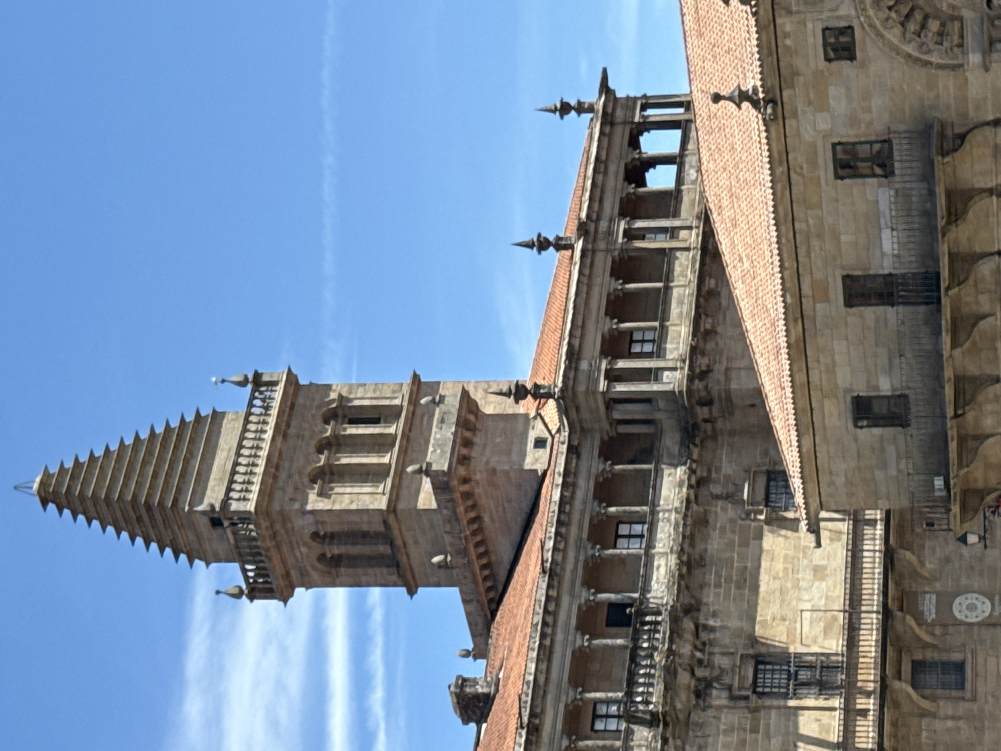 The image depicts a detailed view of the Obradoiro facade of the Santiago de Compostela Cathedral, specifically focusing on its impressive tower and surrounding architectural elements. The scene is bathed in bright sunlight, casting distinct shadows and highlighting the intricate stonework. The sky above is a clear, vibrant blue, with a few wispy white clouds present.
The central focus is a tall, ornate stone tower with a layered, pyramidal spire. This tower exhibits classical architectural features, including arched openings and a decorative balustrade. Extending from the tower, and forming a significant portion of the visible facade, is a multi-story building characterized by a loggia with classical columns and red tile roofing. This structure features numerous windows and balconies, showcasing weathered stone facades.
In the foreground, another section of the cathedral complex is visible, also with red tile roofs and stone construction. This lower building displays similar architectural styles with windows and balconies, contributing to the overall impression of a grand, historic edifice. The stonework throughout the image appears ancient and well-maintained, suggesting a place of historical and architectural significance. There are no people or animals visible in the frame. The overall environment is urban, defined by the monumental architecture of the cathedral.