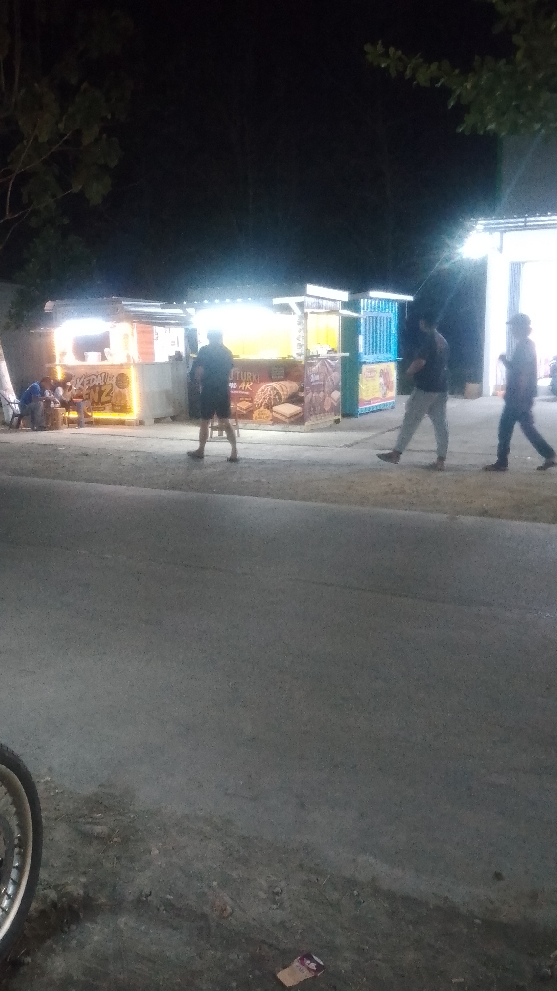In Karanglangu, Indonesia, at night, a street scene unfolds with a row of illuminated food stalls. Three men are visible walking along the pavement in front of the stalls. The stall on the far left is labeled "KEDAI KANZ," while another stall displays images of what appear to be waffles and cookies, with the text "J. TURKI" and "N AK" visible. Further down the row, a bright blue stall stands out. To the far left, two individuals are seated at a table outside one of the stalls. The pavement area in front of the stalls is paved with a rough, grey material, and the road in the foreground is asphalt. In the lower left corner, a portion of a motorcycle wheel is visible. The background is dark, with trees and branches silhouetted against the night sky.