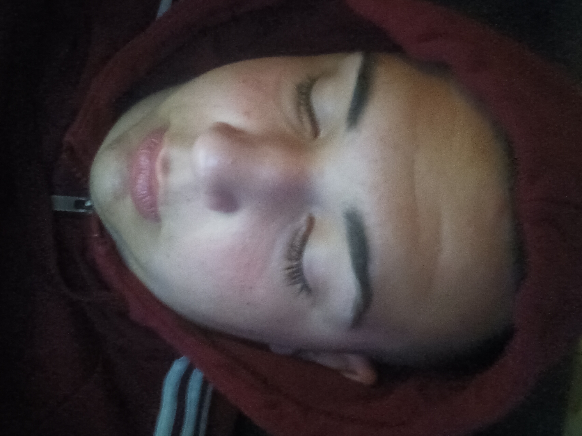 A close-up, angled shot shows a person's face, eyes closed, surrounded by a red hooded garment. The face is pale with visible eyelashes and closed lips. The hood appears to be part of a zippered sweatshirt, and a portion of light-colored stripes is visible on the left side of the frame. The location is Unknown, Unknown.