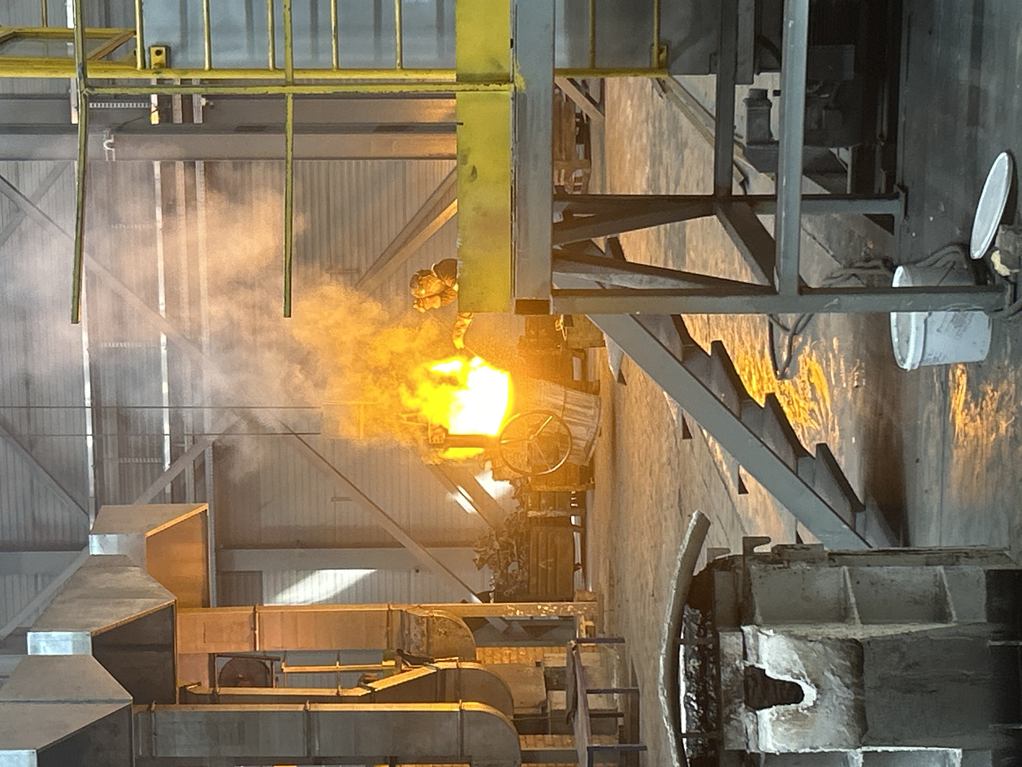 The image depicts an industrial setting, likely a metal foundry. A bright, intense burst of light, possibly molten metal or a fire, is the focal point. Smoke and fumes are visible around the light source. Heavy machinery, ventilation ducts, and metal structures are present. The presence of yellow safety railings and possibly a worker in protective gear suggests human activity is involved. The location in Noviy, Russia, is not directly verifiable from the image alone.