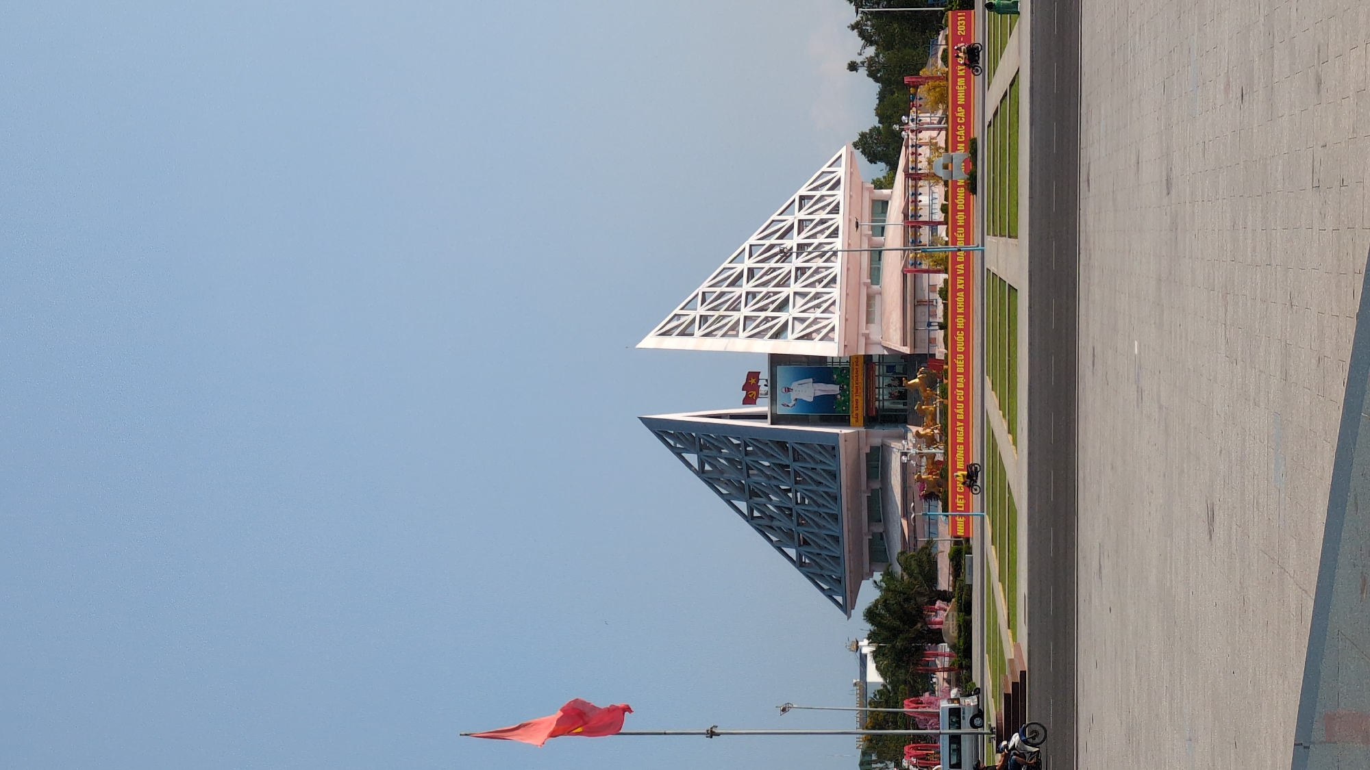 The image shows a view of a public square in the city of Phan Rang – Tháp Chàm, Vietnam, featuring a building with a distinctive architectural design. The building has two triangular structures atop, one white and the other gray, both with a lattice-like framework. A large banner with Vietnamese text is visible in front of the building. A Vietnamese flag waves on a pole to the left. The foreground is occupied by a paved plaza that extends to the right. The sky is a clear blue. Vegetation is visible along the sides of the plaza, including trees and grassy areas. A few parked scooters or motorcycles are along the roadside, and banners are visible along the building front.