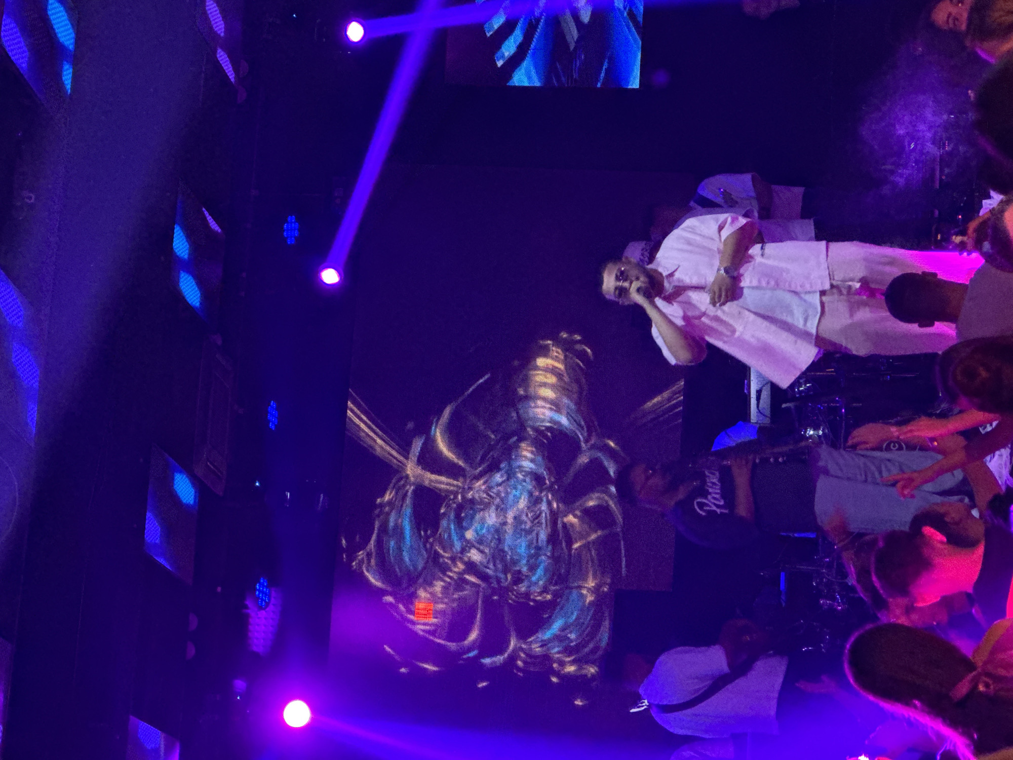 This image depicts a lively concert scene at an indoor venue in Tsarevo, Bulgaria. A male performer, wearing a light pink shirt and matching pants, is center stage, holding a microphone and appearing to sing. He wears sunglasses. Behind him, another male is visible, wearing a dark shirt with the word "Paros" on it, and light-colored pants, holding a musical instrument, possibly a clarinet or saxophone. To the far left, another person in a white shirt with a guitar strap is seen, also appearing to be a musician.
The stage is illuminated by vibrant purple lights and beams. A large screen behind the performers displays abstract, colorful graphics. The foreground shows a crowd of people, many with their hands raised, enjoying the performance. The atmosphere is energetic, indicated by the lighting and the visible audience engagement. The overall environment suggests a nightclub or a concert hall.