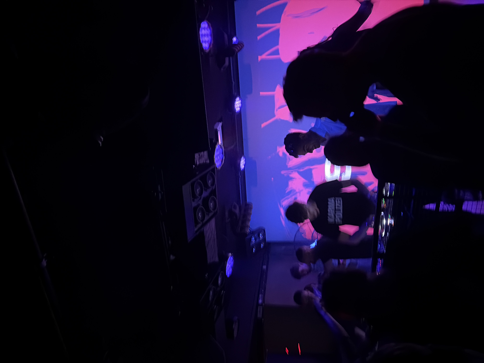 In a dimly lit nightclub with purple and pink lighting, several people are gathered around a DJ booth. A DJ, wearing a black t-shirt with white lettering that reads "KARMA POLIZE," is actively mixing music on a set of turntables. Two other individuals are visible in the background, one appearing to be a man in a light-colored shirt looking towards the DJ, and another person whose features are obscured. The overall atmosphere suggests a lively event, likely a music performance or party. The location is an unspecified nightclub in an unknown city.