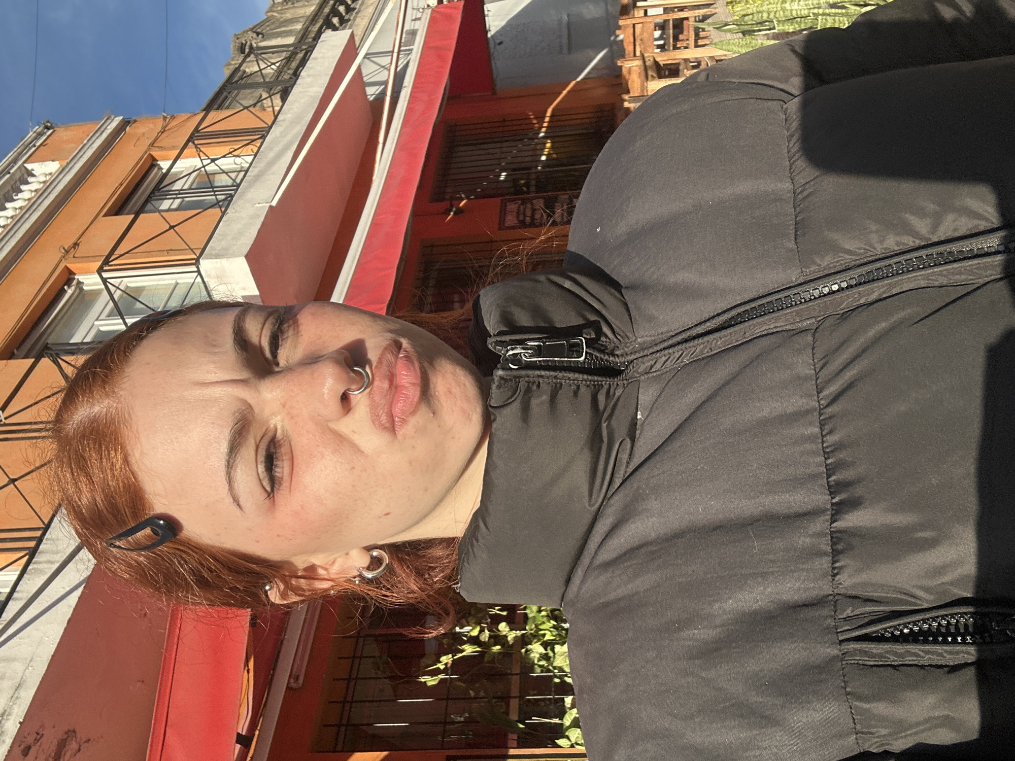 A red-haired individual with freckles and multiple piercings is seen in a medium shot, partially obscuring a street scene. The person wears a black puffer jacket. In the background, there are buildings with orange facades, and a red awning is visible. Vegetation is also present. Based on architectural style and color schemes, this location could be within the city of Buenos Aires, Argentina.