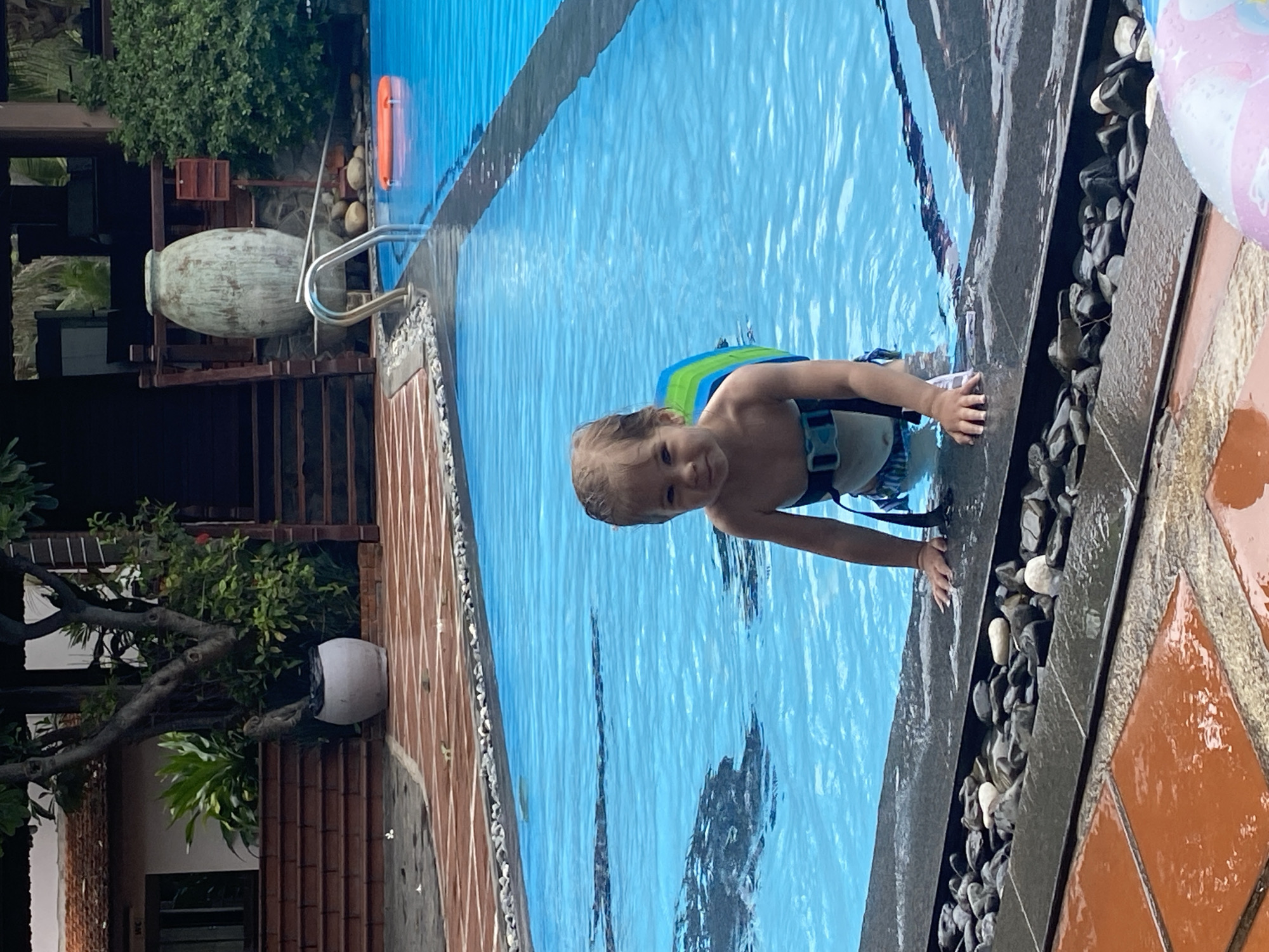 A fair-skinned toddler wearing a blue and green flotation device is in a swimming pool. The pool is edged with dark stones and terracotta tiles. The location is Phu Thuy Ward, Vietnam. In the background, there are buildings, plants, and a decorative urn.