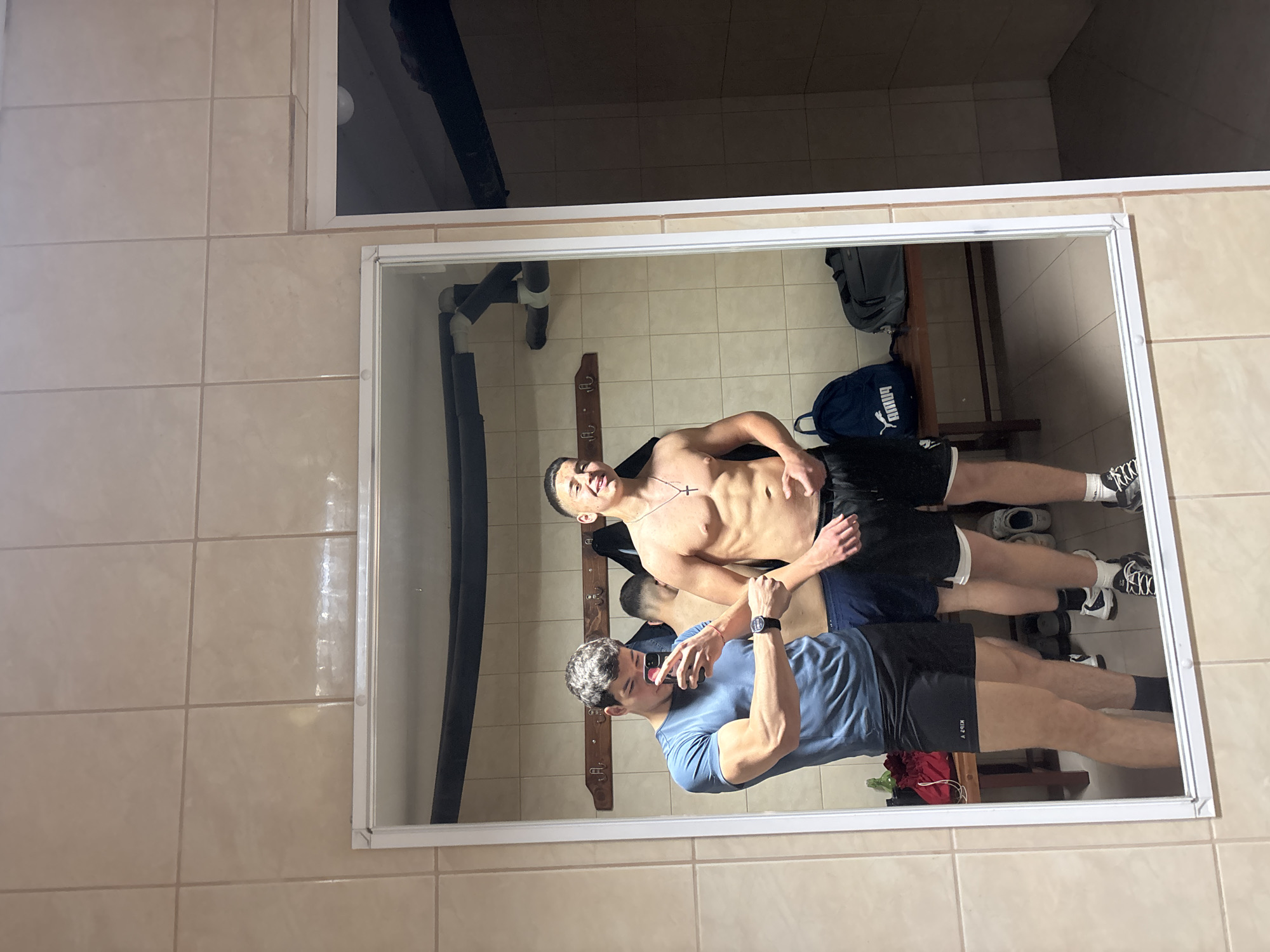 Two young men are reflected in a mirror. The man closest to the viewer is wearing a blue t-shirt and black shorts and holding a phone, presumably taking the picture. The other man is shirtless and wearing black shorts. They appear to be in a locker room. Behind them is a bench, lockers, bags, and shoes. The walls are tiled. The location is potentially Zlatograd, Bulgaria.
