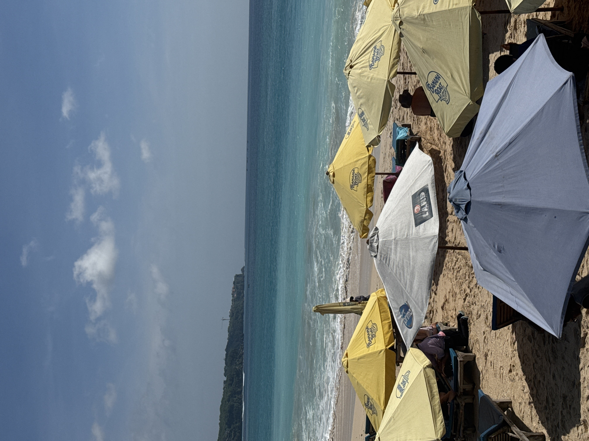 The image shows a beach scene in Kuta Selatan, Indonesia, evidenced by the presence of "Banana Boat" branded parasols. The scene is captured from a high vantage point, presenting an array of beach parasols in the foreground, set against the backdrop of the ocean and coastline. The beach parasols are predominantly yellow and light grey in colour. The beach is sandy, with evidence of beachgoers and their belongings beneath the parasols. The ocean is turquoise in colour, showing small waves breaking on the shoreline. The sky is blue with scattered clouds. The coastline is covered in vegetation, possibly a cliff or a headland.