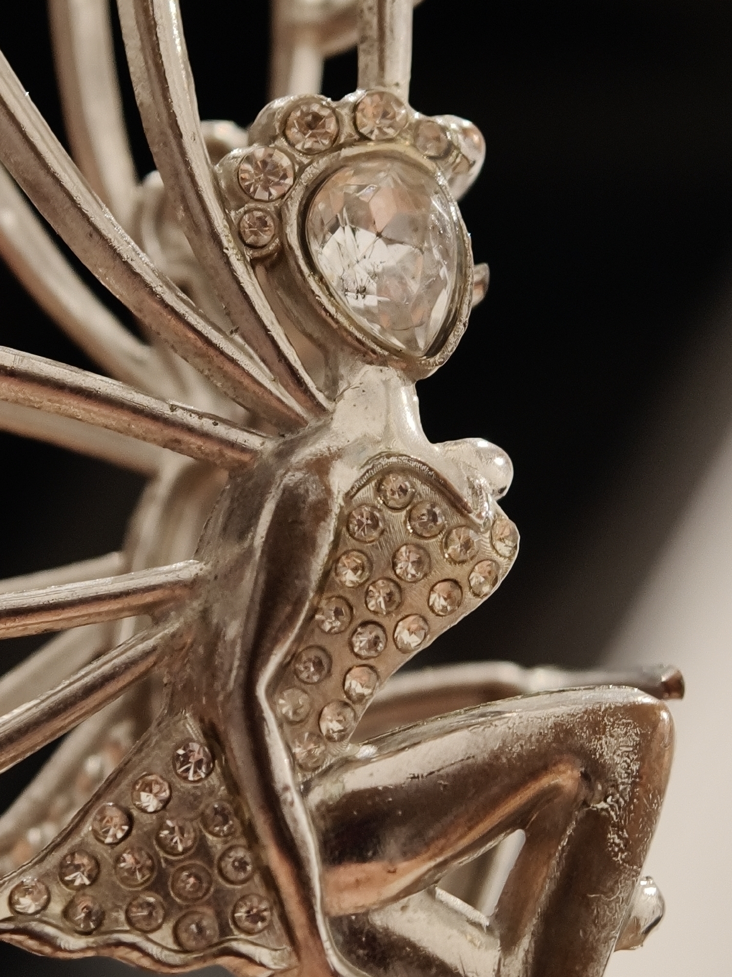 The image is a close-up, macro photograph of a silver-colored fairy figurine adorned with small, clear crystals. The figurine is depicted in a seated position, with one leg bent and the other extended. It has delicate wings fanning out behind it, composed of thin, radiating silver bars. The fairy's head is tilted slightly, and she appears to be wearing a small crown or diadem, also embellished with crystals. The surface of the figurine shows some wear and subtle scuff marks, indicating it is not new. The background is dark and out of focus, with a sliver of lighter color visible on the right side, suggesting a contrasting surface or light source. There is no discernible connection to Đà Nẵng, Vietnam, as the image focuses exclusively on the object itself and lacks any contextual environmental cues.