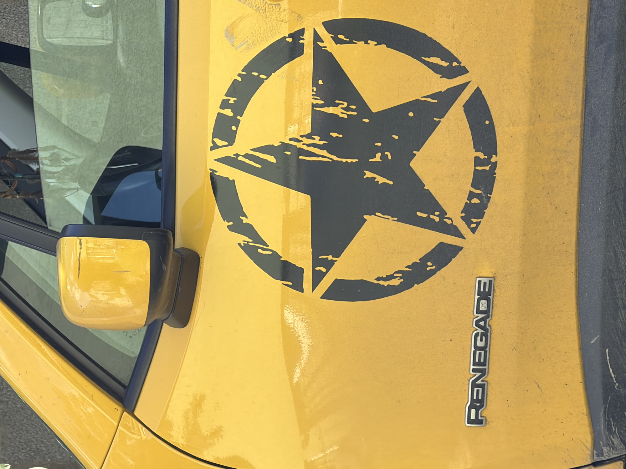 A close-up shot from above shows the hood of a yellow Jeep Renegade, featuring a large, black, distressed star enclosed in a circle. The word "RENEGADE" is displayed vertically in chrome lettering on the hood near the right edge. A portion of the driver's side window and side mirror are visible on the left side of the frame. The scene suggests the vehicle is parked outdoors. While the location is Saguramo, Georgia, specific details tying it to that city are not discernible from the image alone.