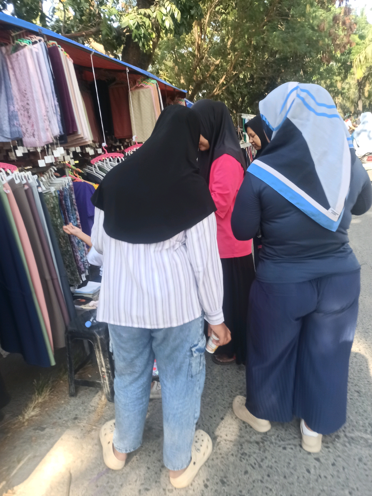 The image shows an outdoor market scene in Telagamurni, Indonesia, with three women visible. The woman in the foreground, facing away from the camera, wears a black hijab, a striped long-sleeved shirt, and light blue jeans with beige clogs. To her left, another woman is partially visible, wearing a black hijab and a pink shirt. To the right of the foreground woman is a third woman, also facing away, wearing a blue long-sleeved shirt, a blue and white headscarf, and dark blue wide-legged pants, with beige clogs. The women appear to be browsing at a stall displaying various clothing items, including what seem to be scarves and fabrics, hung on racks under a blue and orange awning. Trees and foliage are visible in the background. The ground is paved with asphalt.