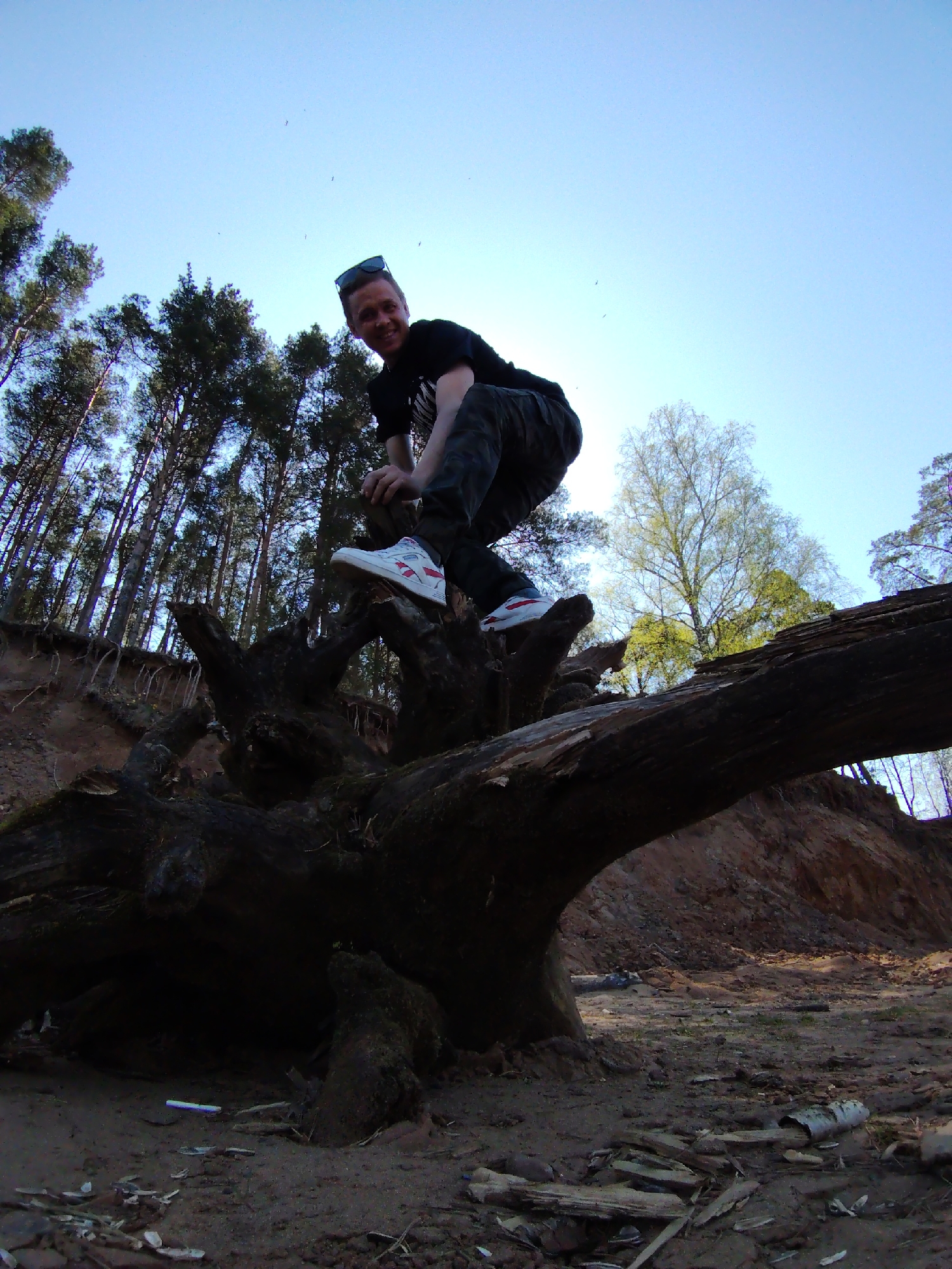 A man is standing on a large, fallen tree trunk in a forest setting. He is wearing a black T-shirt, dark patterned pants, and white sneakers with red accents. Sunglasses are perched on his head. He is looking towards the camera. The tree trunk is thick and moss-covered. In the background, there are tall trees, a sandy bank, and a clear blue sky. The ground is covered in mud, small rocks, and scattered twigs. This location is in the city of Chaikovsky, Russia.