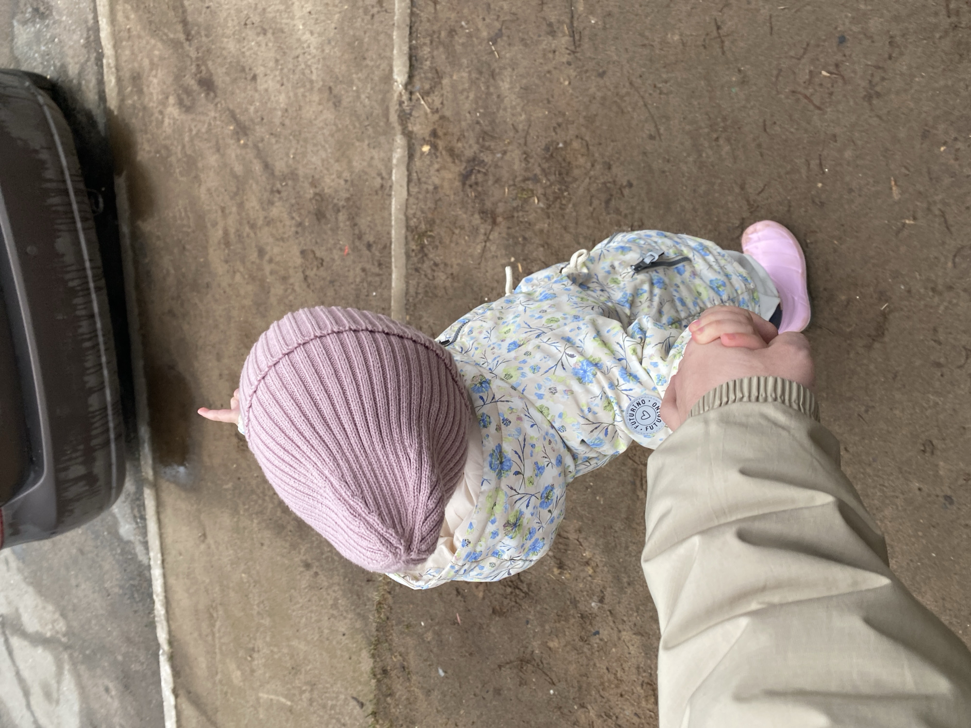 The image, taken from a slightly elevated, overhead perspective, captures a small child being guided by an adult outdoors in Chaikovsky, Russia. The scene is set on a mottled, damp-looking concrete or paved surface, with what appears to be the tire of a dark vehicle visible on the far left.
The child, seemingly a toddler, is dressed in warm attire. They wear a ribbed, light purple or mauve knit beanie on their head. Over their clothes, they have on a light-colored jacket or dress adorned with a floral pattern of blue and light green leaves and flowers. On the child's right wrist, a circular tag is visible, displaying the text "ON FUTURINO" and partially "FUTURINO". Their feet are clad in bright pink, possibly rubber, boots. The child's left hand is held firmly by an adult's right hand.
Only the arm and hand of the adult are visible, extending from the right side of the frame towards the child. The adult wears a light beige or khaki-colored jacket with a slightly wrinkled texture and a ribbed cuff at the wrist.
The ground beneath them is a mix of concrete and possibly dirt, showing signs of dampness or recent rain. The overall lighting suggests an overcast or cloudy day. The image captures a moment of an adult holding a child's hand, presumably walking or standing together.