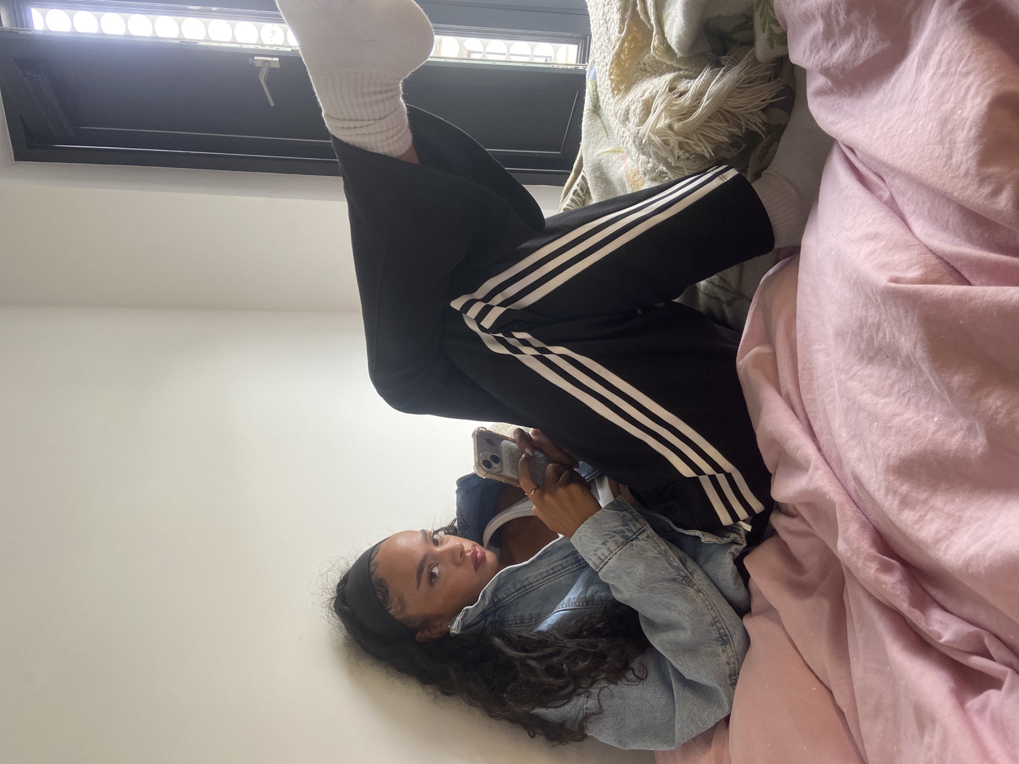 This rotated image captures a young woman lying on her back, her head towards the left side of the frame, and her legs elevated towards the top. She has long, dark, curly hair held back by a black headband. She is wearing a light blue denim jacket over a white top, black Adidas track pants with three white stripes along the sides, and white ribbed socks on her elevated feet.
The woman holds a silver smartphone in her hands, its screen facing her, and she appears to be looking towards the camera with a calm expression and slightly parted lips. She is positioned on a bed, with a white wall behind her head and a textured cream-colored blanket or throw visible near her raised legs. To her right, a voluminous pink duvet or blanket is crumpled, adding a soft texture to the scene.
At the top of the frame, a dark-framed rectangular window or skylight is visible, allowing bright light into the room and showing multiple circular patterns within its structure. The overall setting is indoors, likely a bedroom, characterized by simple white walls and comfortable bedding. The light suggests it is daytime. There are no specific location cues visible within the image to indicate London, United Kingdom, and no visible text. The atmosphere appears relaxed and casual.
