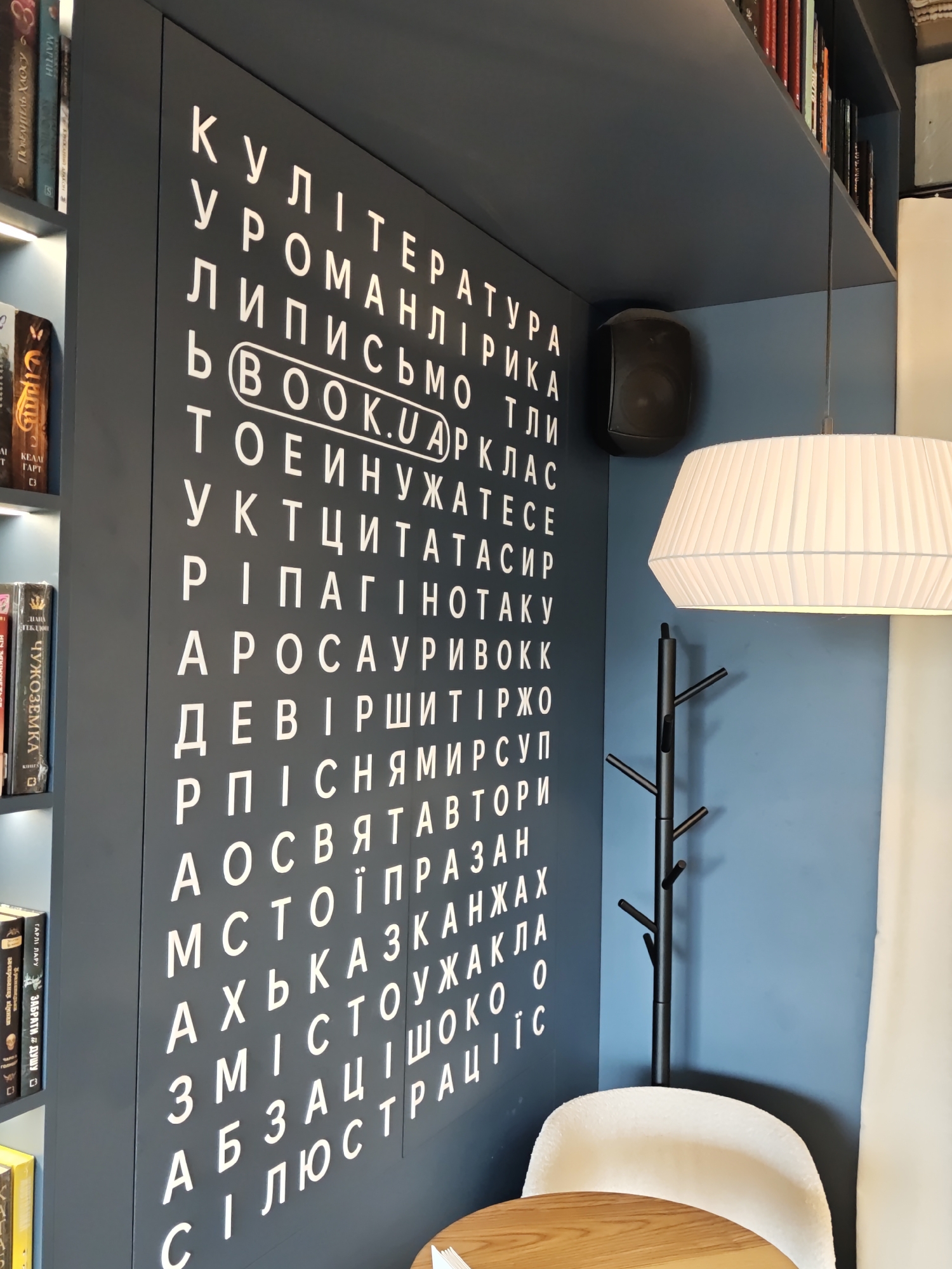 The image shows a wall with white letters in Ukrainian arranged in a grid, resembling a word search puzzle. The wall is painted dark blue. A circular logo with "BOOK.UA" inside is highlighted in the grid. To the left, a bookshelf is partially visible with books. To the right, a white lampshade hangs from the ceiling, and a black coat rack stands near a white chair. The overall impression is of a cozy, possibly a bookstore or cafe interior. The inscription and the logo suggest the location is likely related to books and the city of Kyiv, Ukraine.