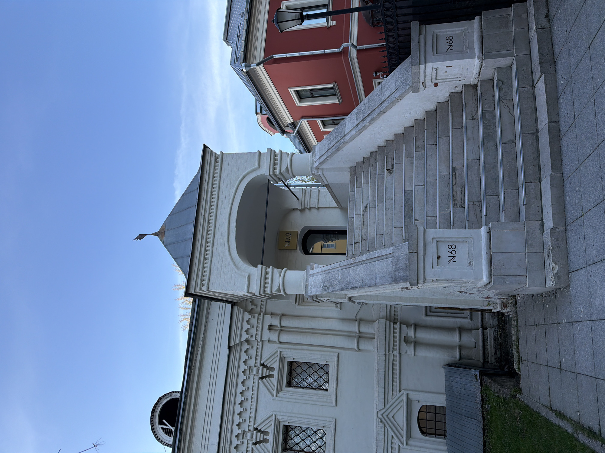 The image captures an architectural scene in Moscow, Russia, under a clear blue sky during the daytime.
**Subjects:** The primary subject is a grand, historic white building, possibly a church or a palace, distinguished by its ornate architectural details. These include decorative moldings, pilasters, and small rectangular windows fitted with dark metal grilles. A large arched entranceway is visible on its side, beneath a dark, possibly gray, roof or dome that culminates in a small finial. To the rear and right of the white building, a red-colored building with white window trim and a dark roof is visible, featuring a traditional black street lamp and part of a decorative black fence. A substantial stone staircase leads up to the entrance of the white building.
**Activity or Event:** No specific activity or event is taking place; the image presents a static view of the architectural structures.
**Notable Details:** The time of day is clearly daytime, indicated by the bright, clear blue sky with only a minimal, wispy cloud. The weather appears fair and sunny. The architectural style of the white building is historic, featuring intricate stone carvings and details. The stone staircase includes a sturdy balustrade and is made of light gray, possibly limestone, blocks. The ground beside the staircase is paved with similar gray tiles, with a small patch of green grass visible at the base of the white building.
**Visible Text:** The alphanumeric text "N68" is visible in multiple locations. It appears prominently on a square plaque on the stone balustrade of the staircase, engraved or painted. A similar "N68" is also seen on a smaller, golden plaque positioned above the arched entrance of the white building, and again on a stone block near the base of the staircase.