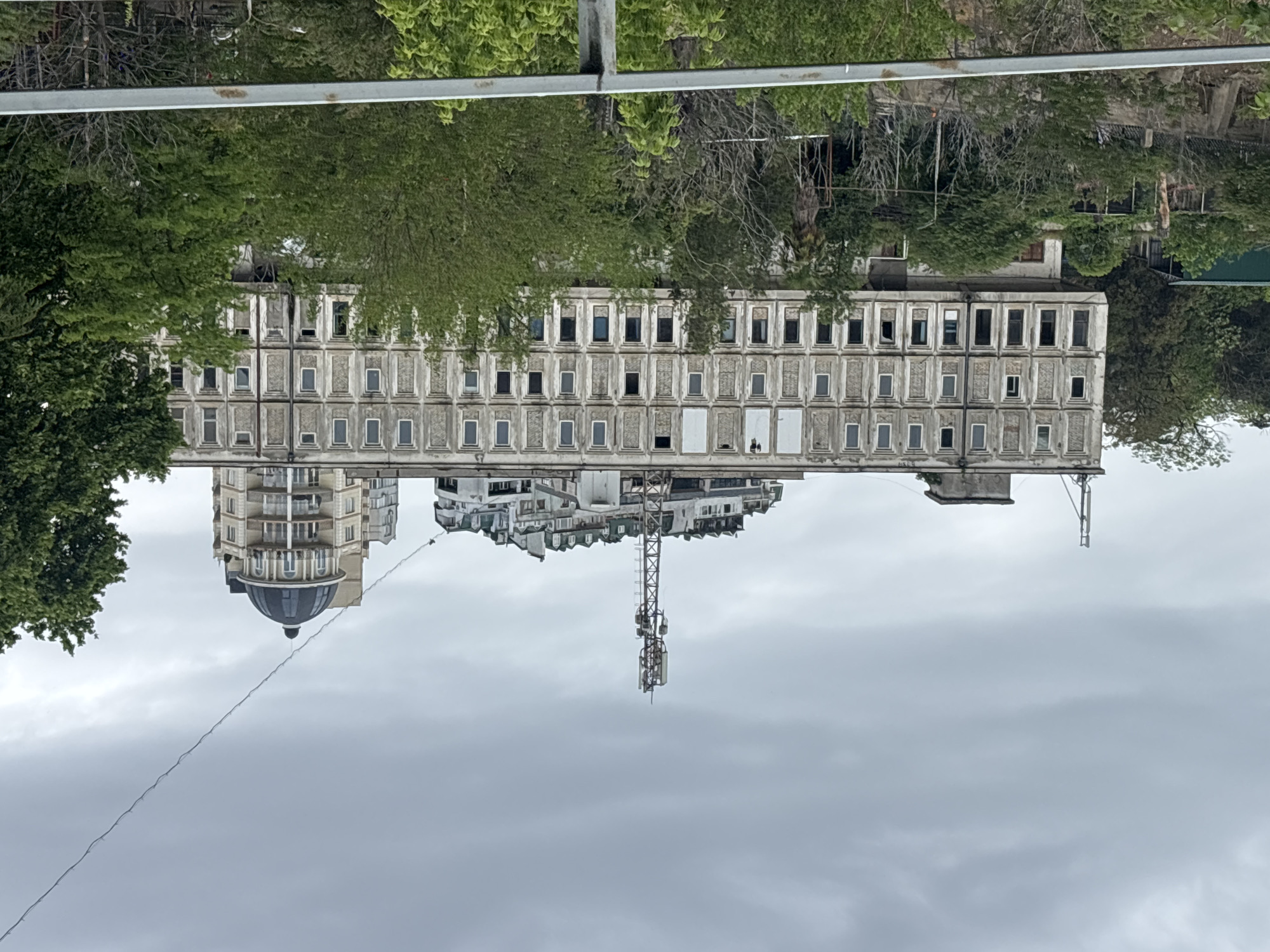 The image shows a building complex in Tbilisi, Georgia, taken from a ground-level vantage point. The central structure is a long, multi-story building of Soviet-era architecture, with rows of rectangular window openings. Many windows are boarded up or missing. The building's facade is deteriorating. Adjacent to this main building is a rounded structure with a dome. A cellular tower rises from the area behind the Soviet building. The buildings are surrounded by trees and vegetation. The sky is overcast. A metal pole is in the upper portion of the image.