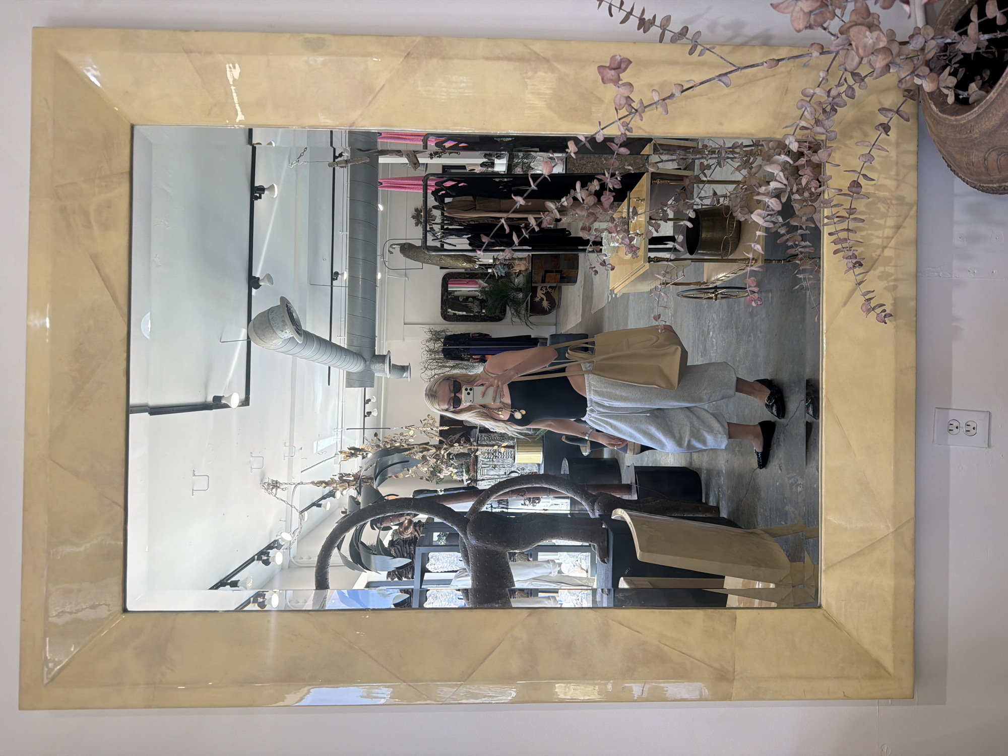 Here's a forensic description of the image:
The image shows a vertically oriented mirror in a retail environment. The mirror has a wide, light-colored frame with an angular, geometric design. The reflection in the mirror reveals a woman holding a cell phone, presumably taking the picture. She is wearing a black top, light-colored pants, and sunglasses. She is also holding a tan tote bag.
The reflected environment appears to be a clothing boutique with racks of clothes, mannequins, and various decorative items. The ceiling has exposed ductwork and lighting fixtures. To the right of the mirror, dried floral branches are visible in a pot, and a standard US electrical outlet is on the wall.
Based on visual clues within the reflection, this is likely a boutique located in Los Angeles, United States of America.