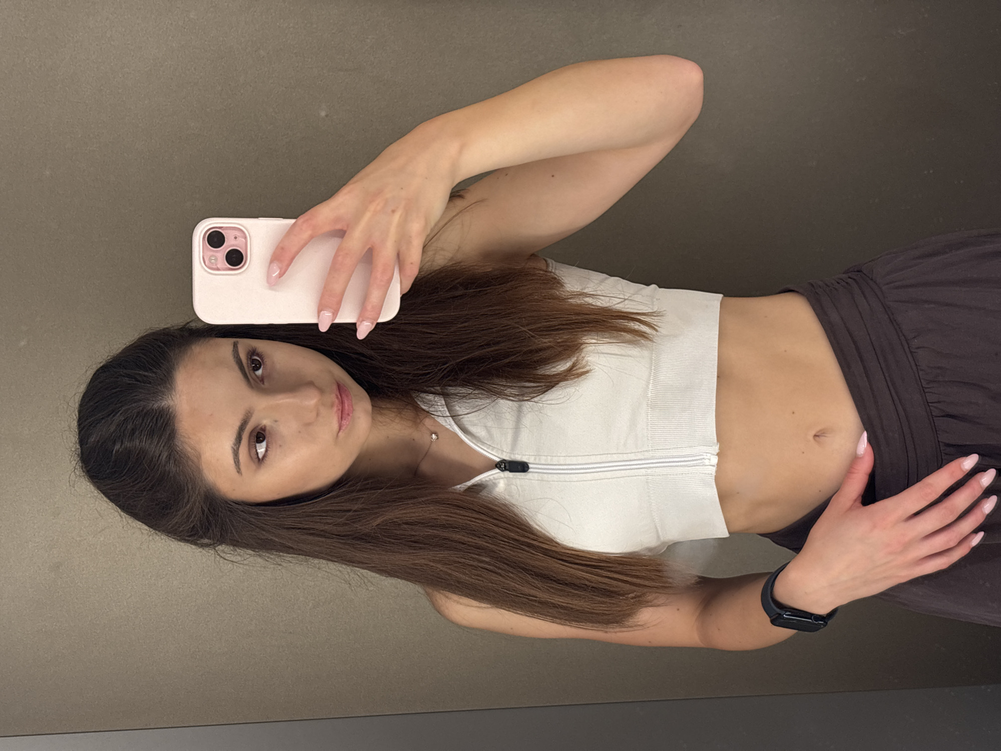 A young woman with long, brown hair takes a mirror selfie while wearing a white zip-up athletic crop top and dark grey workout pants. She has on a black smartwatch on her left wrist and a delicate necklace. Her fingernails are painted a light pink color. She is holding a pink phone in her right hand, angled to capture her reflection. The background appears to be a room with neutral-colored walls. The lighting suggests it is daytime. The image is oriented vertically.
