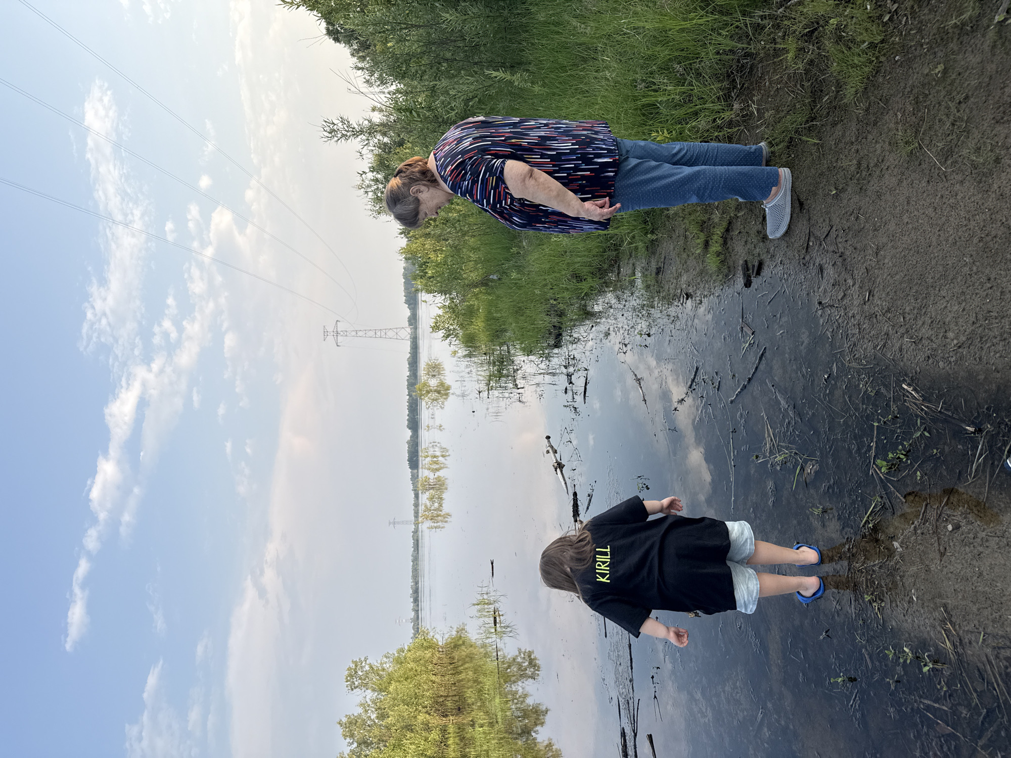 This image depicts two individuals, an adult and a child, standing near a body of water in Barsovo, Russia. The adult, viewed from the side, wears a colorful, patterned shirt and blue jeans, with grey clogs on their feet. They are bent over, looking down at the water. The child, facing away from the camera, is wearing a black t-shirt with the word "KIRILL" in yellow letters on the back, and light-colored shorts. They are also wearing blue flip-flops. Both are standing at the edge of the water, which appears to be a lake or river, with reflections of the sky and surrounding trees visible on its surface.
The scene is set outdoors, with lush green grass and vegetation bordering the water's edge. In the background, a power transmission tower and power lines are visible, along with trees reflected in the water. The sky is partly cloudy, suggesting it could be daytime, possibly late afternoon or early evening, given the soft lighting. The overall mood appears relaxed and natural, with the subjects engaged with the environment. The muddy shore indicates a natural, somewhat undeveloped setting.