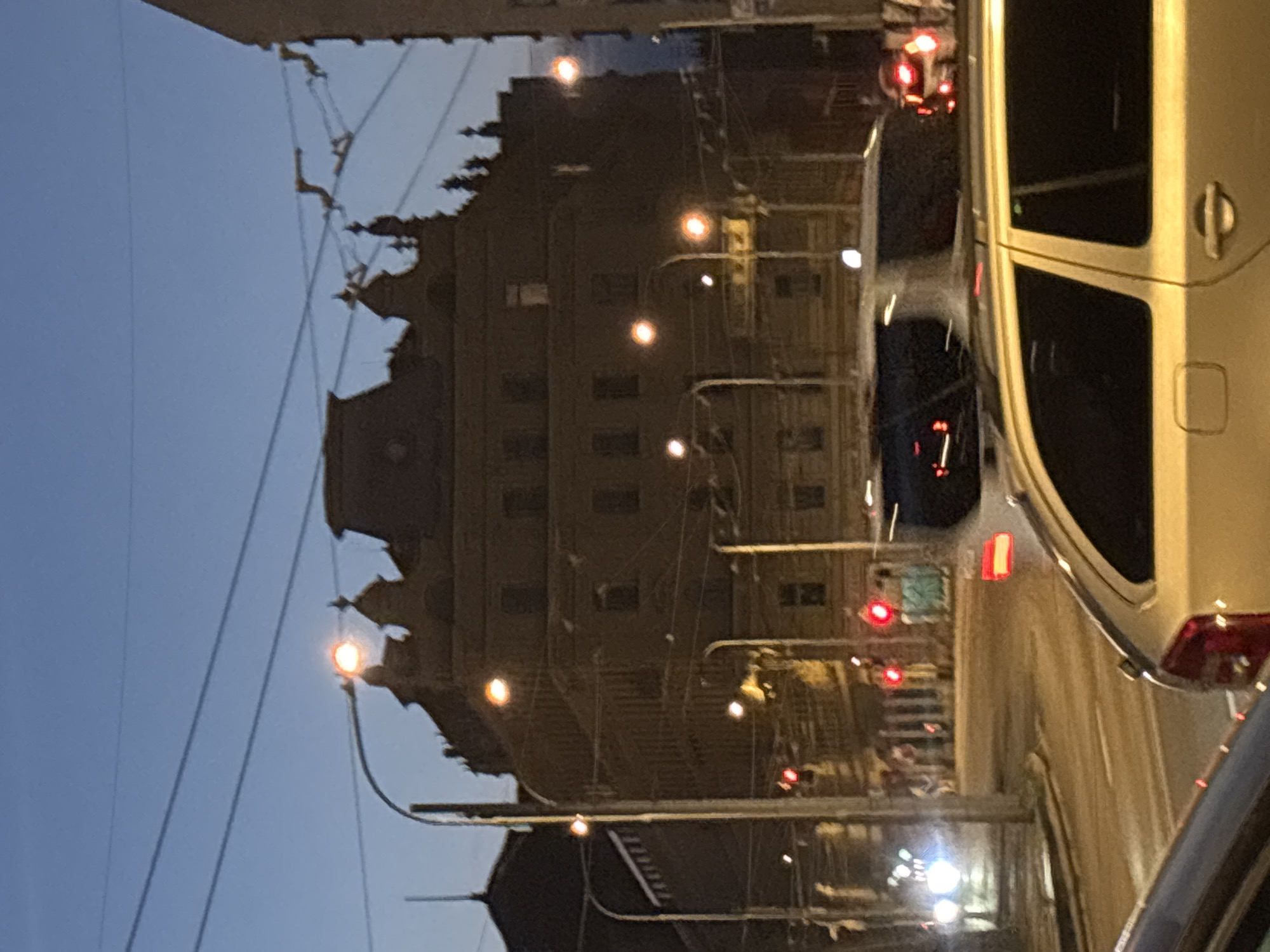 In the city of Unknown, Unknown, an urban street scene is depicted at dusk. A large, dark building with intricate architectural details, including a mansard roof and decorative elements, dominates the background. Multiple streetlights with glowing orange bulbs are visible, illuminating the scene. Overhead, power lines crisscross the sky, suggesting an active urban infrastructure.
In the foreground, several vehicles are present. A gold station wagon is prominently featured, its side profile showing door handles and a rear window. To its left, another vehicle is partially visible. In the middle ground, a white van or bus is seen, along with other cars. Traffic lights are illuminated, with at least two showing red signals, indicating ongoing traffic flow. The street surface appears wet, reflecting the lights and suggesting recent rain or damp conditions. The overall atmosphere is that of a bustling city transitioning from day to night.