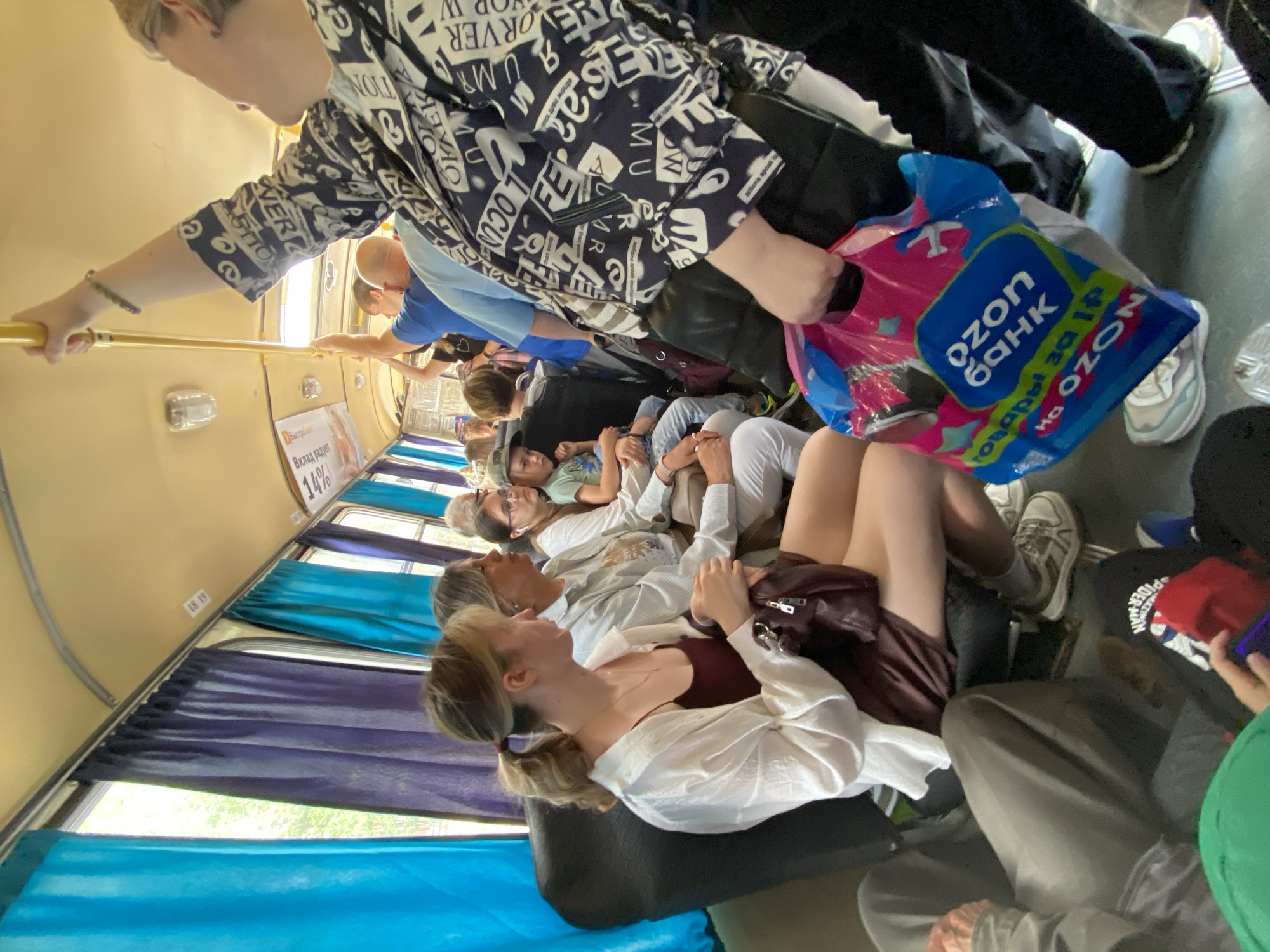 This media file captures an interior scene, likely on a crowded mode of transportation, possibly a bus or train, given the presence of multiple individuals seated and standing, along with overhead luggage racks and curtains. The lighting suggests it is daytime.
The primary subjects are people, a mix of ages and genders, appearing to be passengers. Some are seated, while others are standing, holding onto railings. One person in the foreground is holding a pink and blue plastic bag with "ozon банк" and "товары за 19" visible text. Another individual wears a black and white patterned shirt with text, including "VER" and "TION".
The scene appears to be in motion, with people settled in for a journey. The overall atmosphere seems somewhat cramped and casual.
Visible text includes "18 19" on a wall, a sign that reads "Вклад радует 14%" with the logo "Быстроване", and the branding on the plastic bag and the patterned shirt. The presence of Russian text suggests the location is likely Russia.